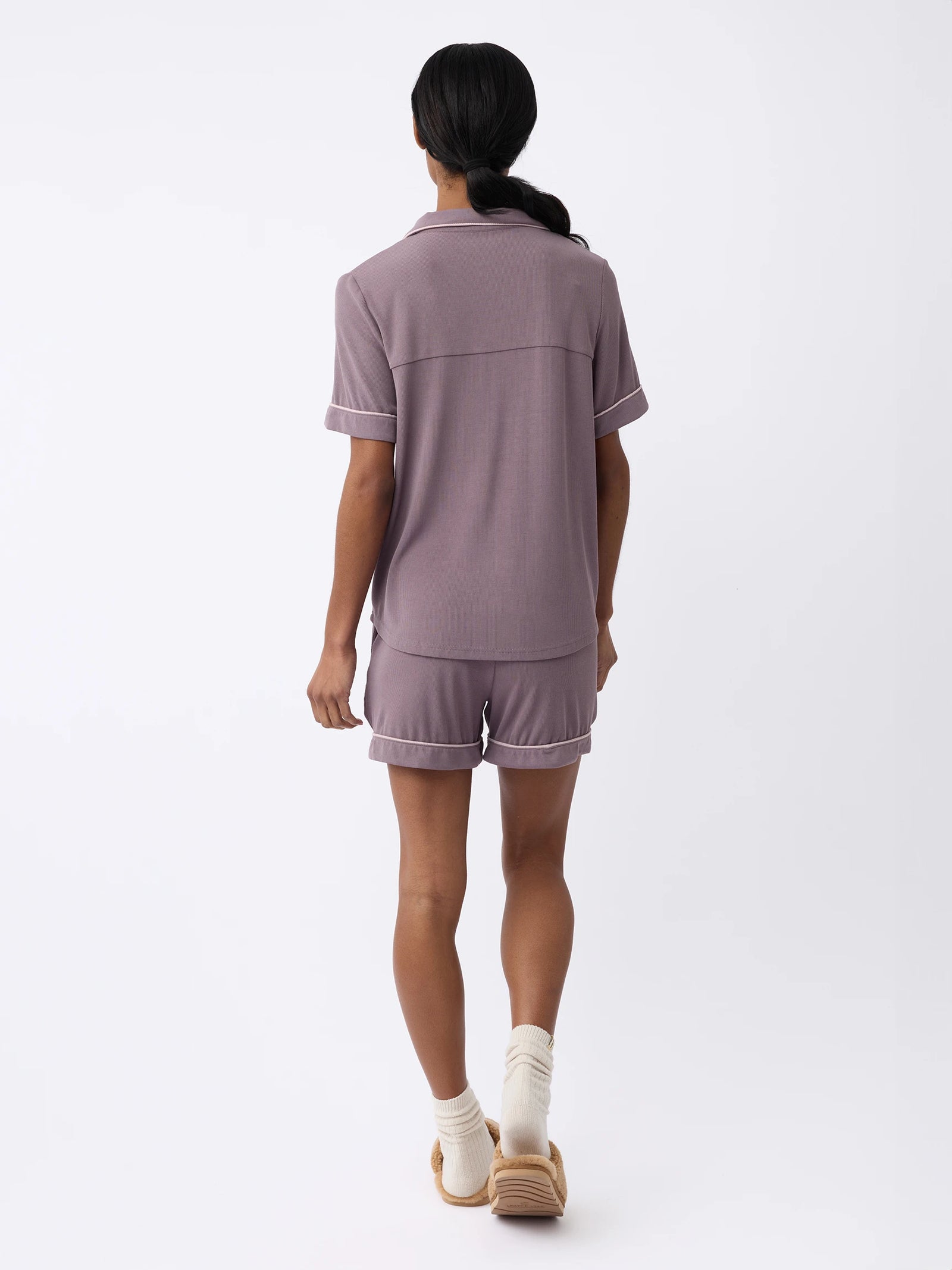 A woman with long black hair is seen from the back, wearing a Cozy Earth Women's Bamboo Rib-Knit Classic Short Sleeve Pajama Top in Twilight and matching shorts. They walk on a white background with white socks and tan slippers. 