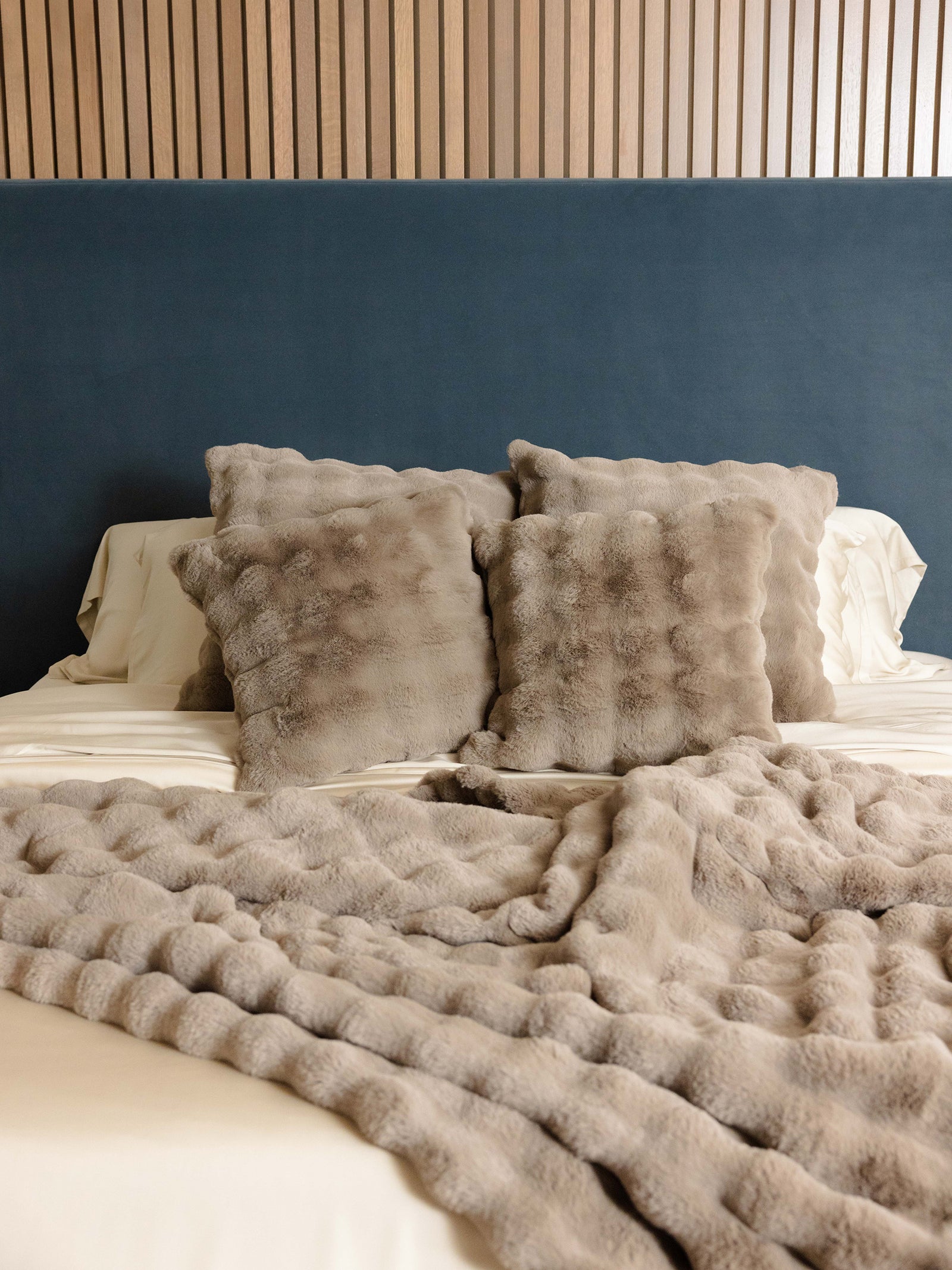 A cozy bed featuring a blue headboard is styled with the Bubble Cuddle Pillow by Cozy Earth, which comes in textured gray, along with a matching gray throw blanket. The wall is adorned with vertical wooden slats, infusing warmth and texture into the bedroom decor. 