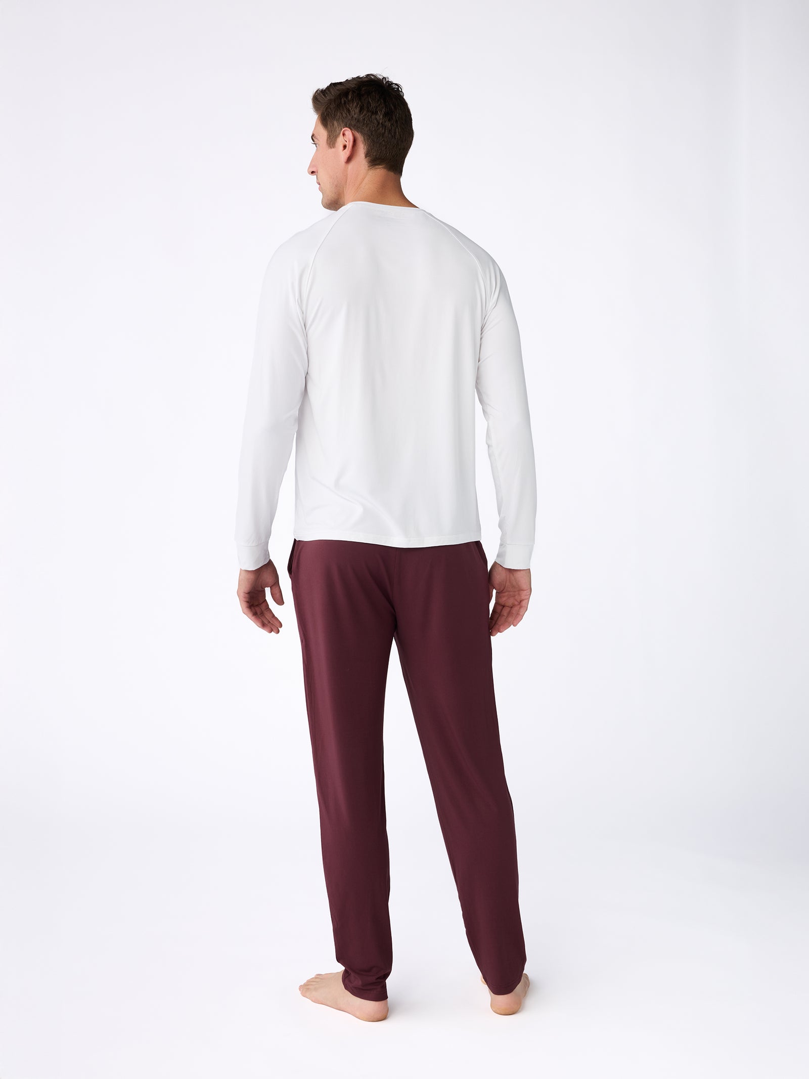 A man stands barefoot on a white background, facing away. He is wearing a long-sleeved top from the Cozy Earth Men's Stretch-Knit Bamboo Long Sleeve collection and maroon pajama pants. 