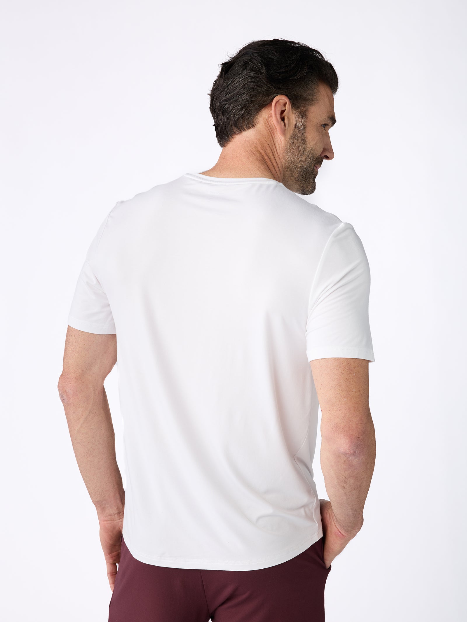 A man with short, dark hair is facing away, wearing a white Men's Stretch-Knit Bamboo Lounge Tee by Cozy Earth and dark red shorts. He stands against a plain, light background. 