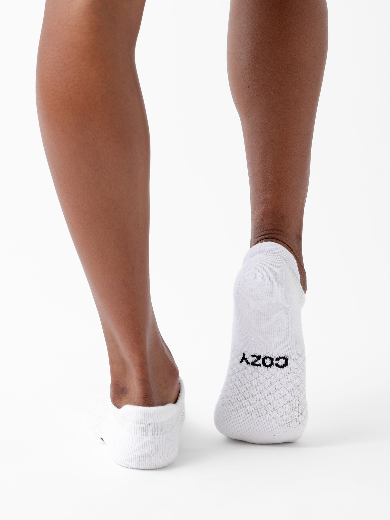 Close-up view of a person's lower legs and feet wearing white ankle socks from the Essential Ankle Sock 2-Pack by Cozy Earth, featuring the word "COZY" written in black on one of the soles. The background is plain white. 