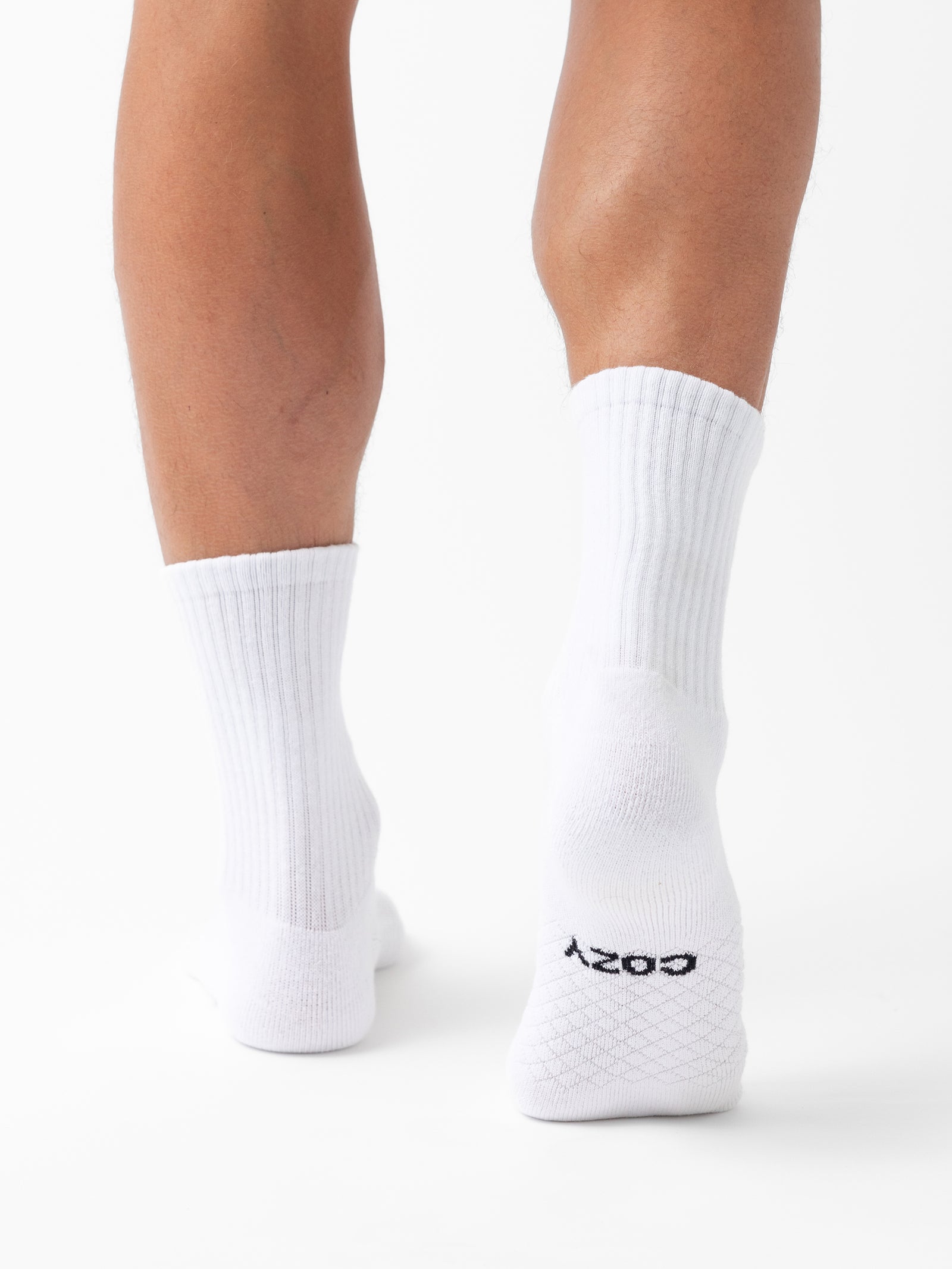 Close-up of the back of a person's legs wearing white ribbed socks with the brand name "Cozy Earth" printed in black on the soles. The background is a plain white surface, showcasing the Essential Calf Sock 4-Pack. 