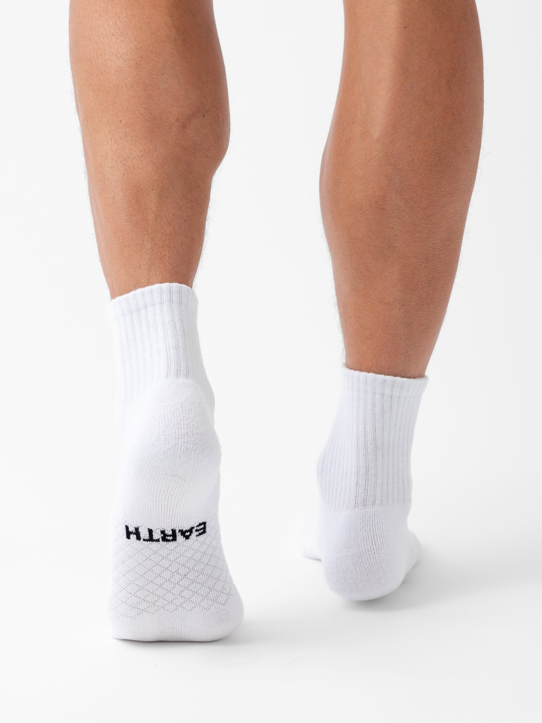 A person wearing Cozy Earth Essential Quarter Socks stands on a white background, back of legs visible. The word "EARTH" is printed in black near the heel on the sole of the left sock. 