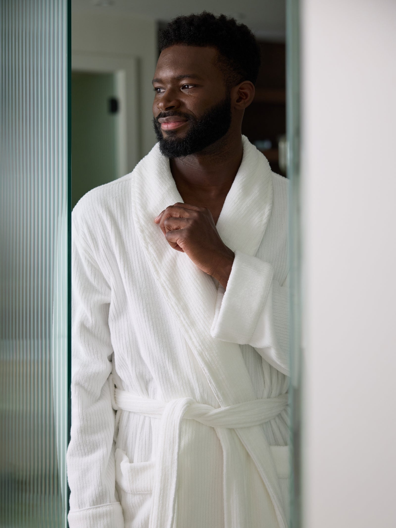 A bearded man stands near a doorway, wearing a Cozy Earth Ribbed Terry Bath Robe in white. He gazes to the side with a slight smile, while the softly lit and blurred background emphasizes his presence. 