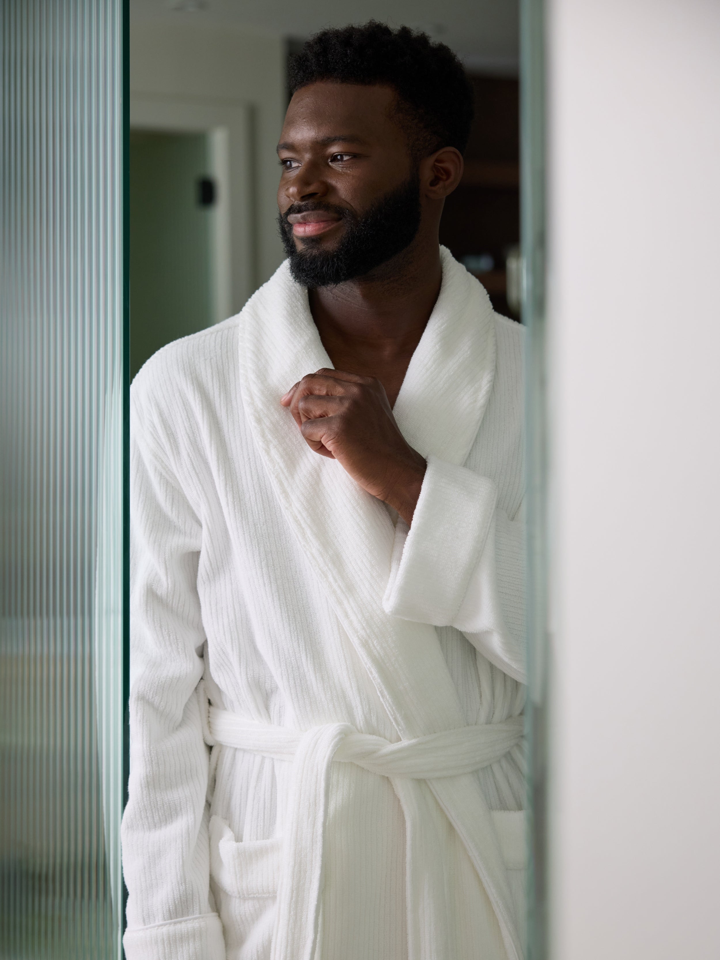 A bearded man stands near a doorway, wearing a Cozy Earth Ribbed Terry Bath Robe in white. He gazes to the side with a slight smile, while the softly lit and blurred background emphasizes his presence. |Color:White