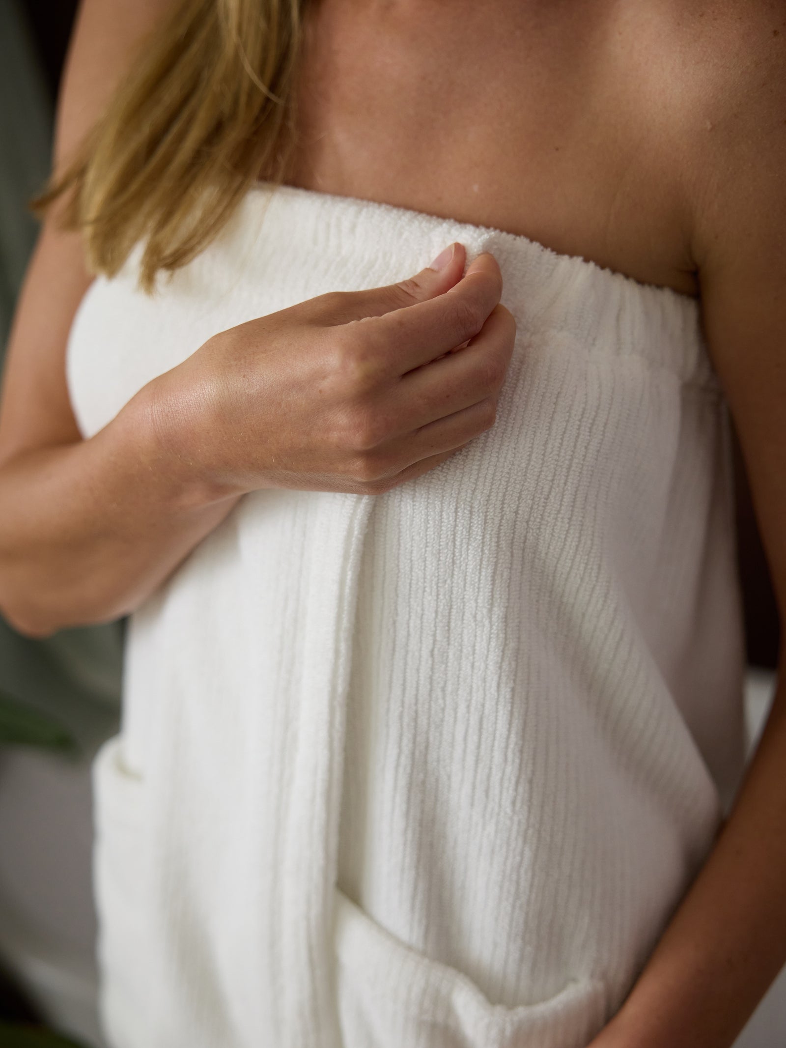 A person with light brown hair is seen from the shoulders down, wearing a strapless Ribbed Terry Bath Wrap by Cozy Earth. They are holding the top edge of the wrap with one hand to keep it secure. The background is blurred and includes shades of green and brown. 