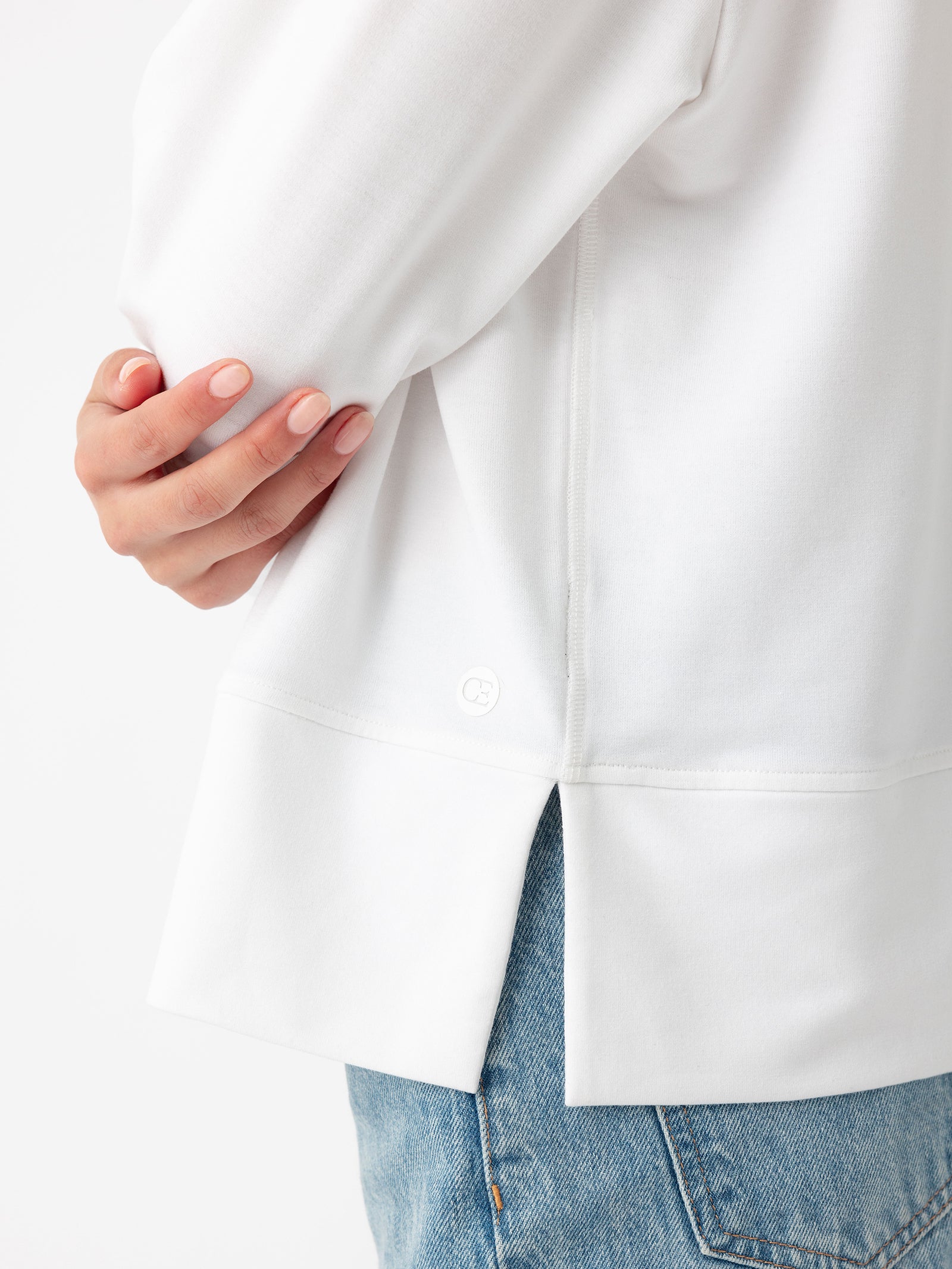 A person is shown from the shoulders down, wearing a Women’s StudioLite Crewneck by Cozy Earth paired with blue jeans. The individual has their right arm gently crossed over their body, resting their hand on the left side. The bottom hem of the shirt features small side slits. 