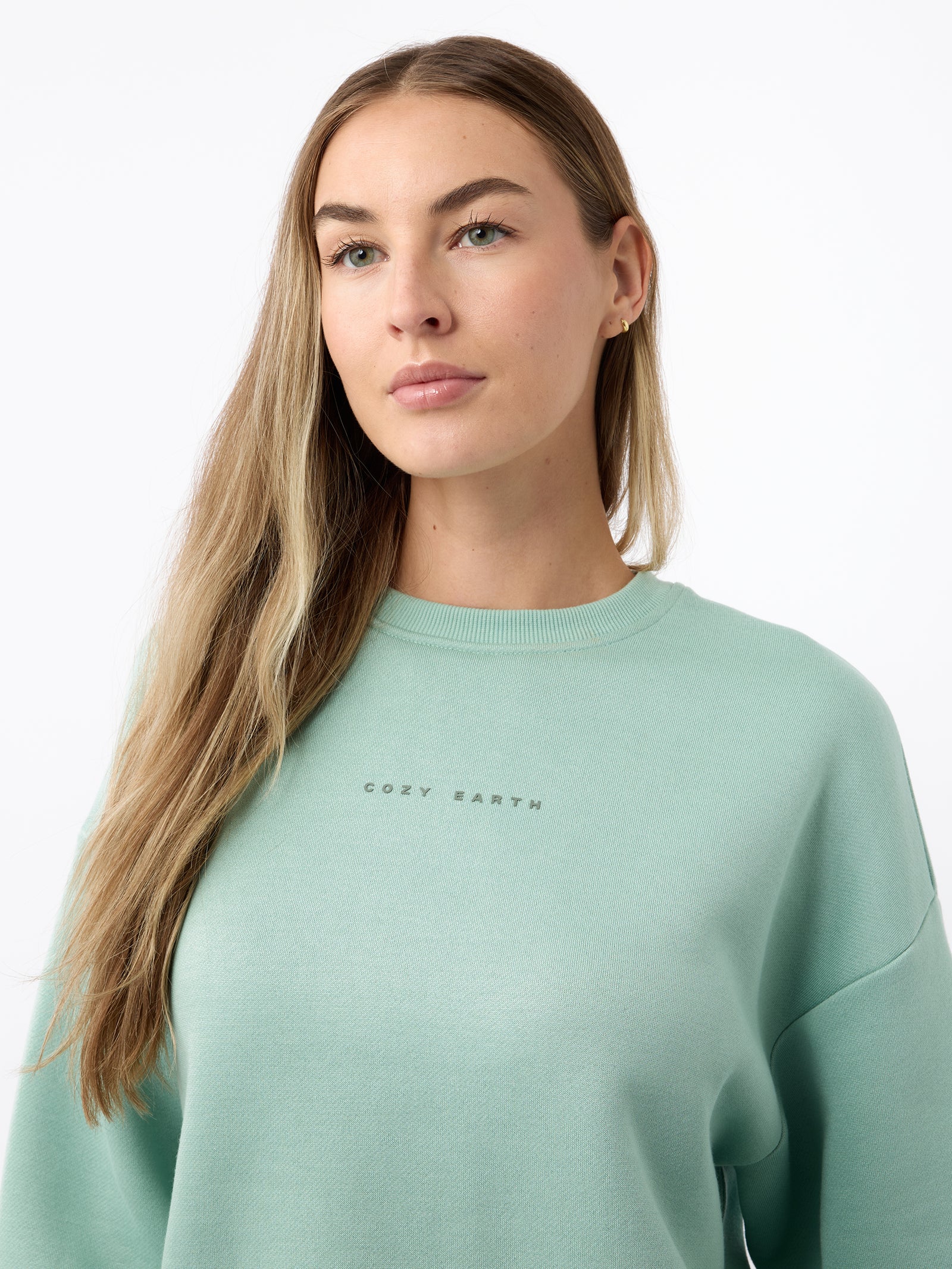 A person with long hair wearing a light green Women's CityScape Crewneck from Cozy Earth stands against a plain white background. 