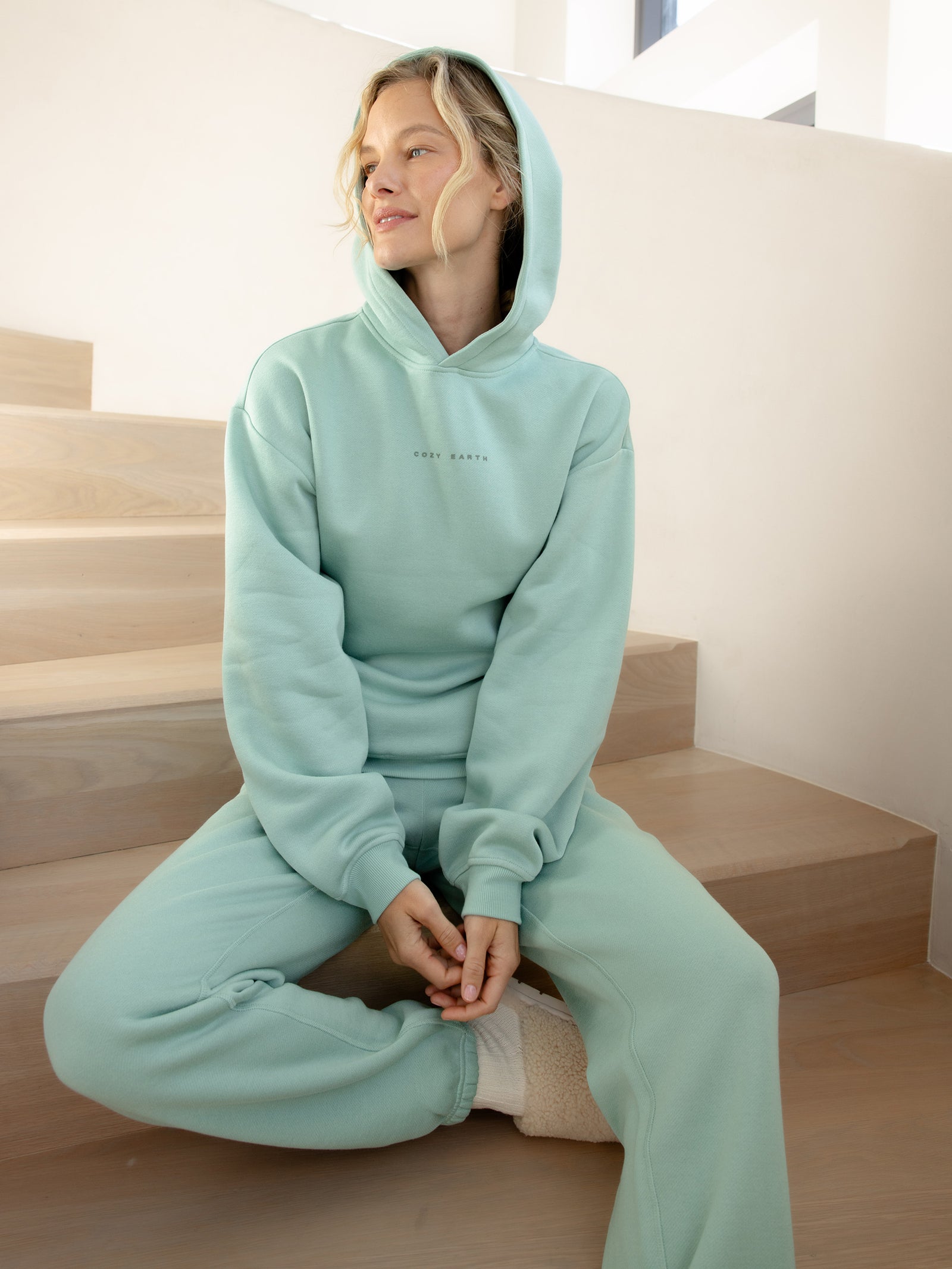 A person sits on wooden stairs wearing a mint green Cozy Earth Women's CityScape Hoodie and sweatpants. They have light hair and are smiling while looking to the side. Soft natural light illuminates the scene, creating a cozy atmosphere. 