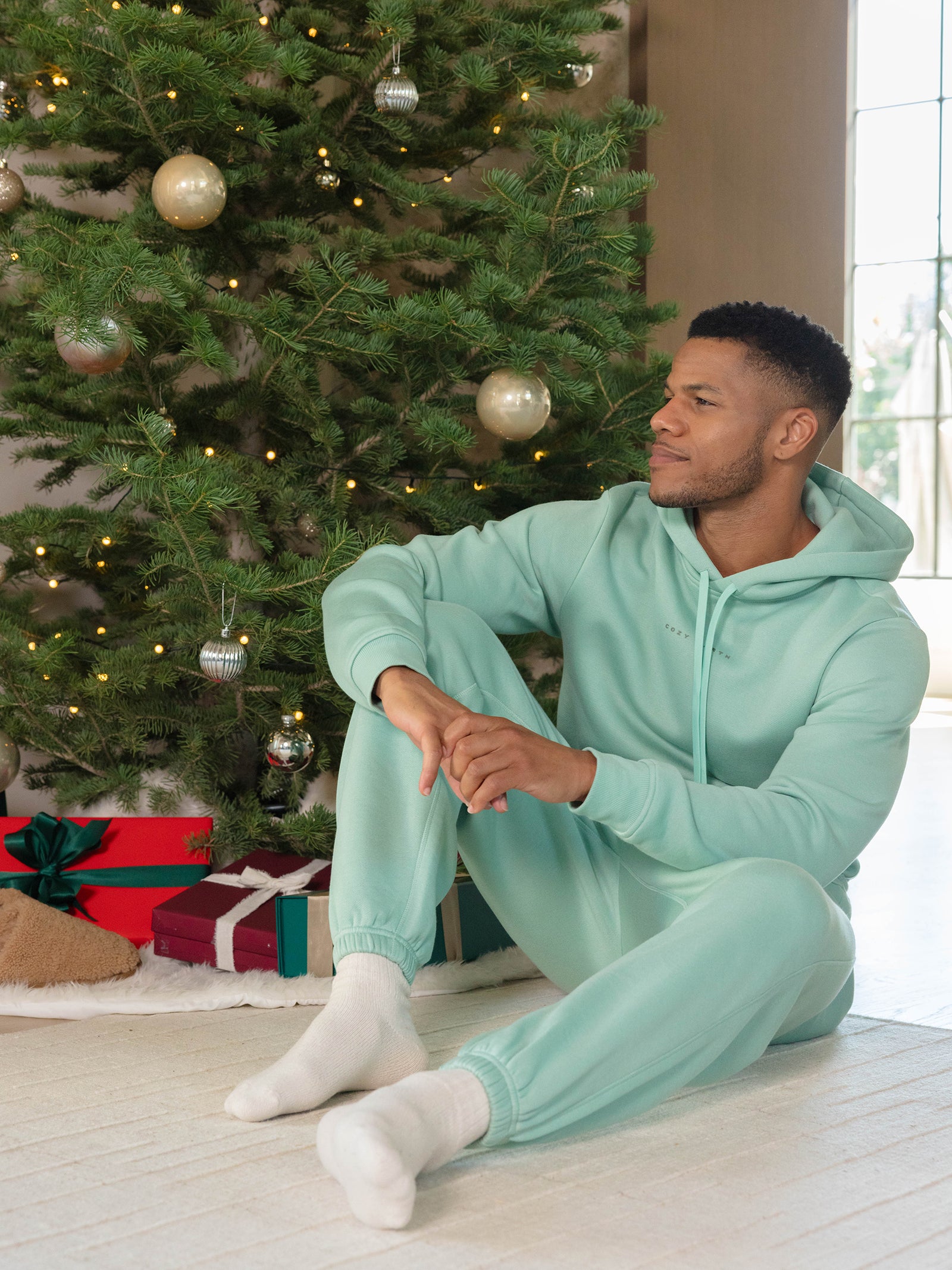 A man wearing a Cozy Earth's Men's CityScape hoodie in teal sits on the floor by a Christmas tree adorned with silver and gold ornaments. Several wrapped gifts are placed under the tree, as soft light filters through a nearby window. 