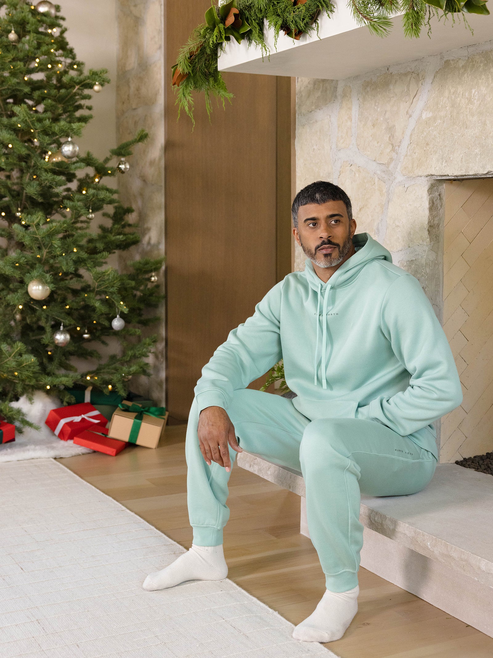 A man in a mint-green CityScape Jogger by Cozy Earth sits by a stone fireplace adorned with greenery. A decorated Christmas tree and wrapped gifts are nearby, creating a festive atmosphere. The room has a cozy and relaxed holiday vibe. 