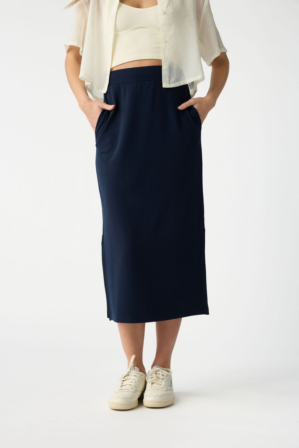 A person wears a white short-sleeve top paired with Cozy Earth's navy Women's Brushed Bamboo Midi Skirt, hands in pockets. White sneakers complete the outfit against a plain white backdrop. |Color:Navy