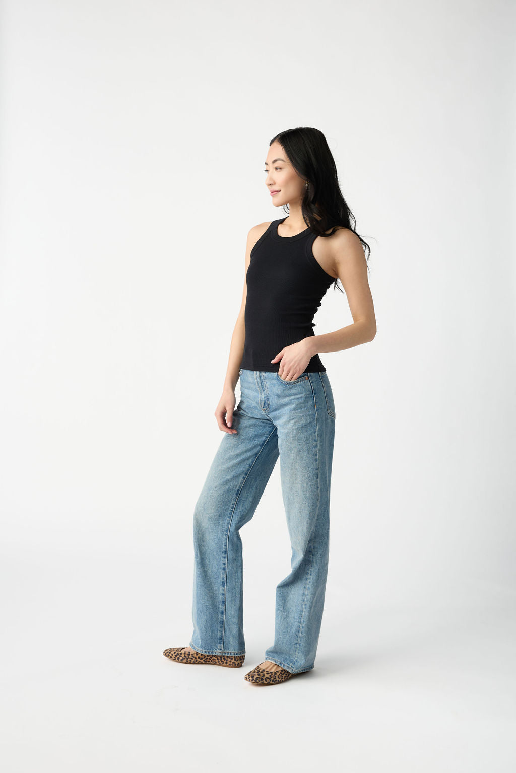 A dark haired woman wears Cozy Earth Women's Fine Ribbed Tank in Jet Black. She puts one hand in the pocket of her blue jeans and stands in front of a white background. 