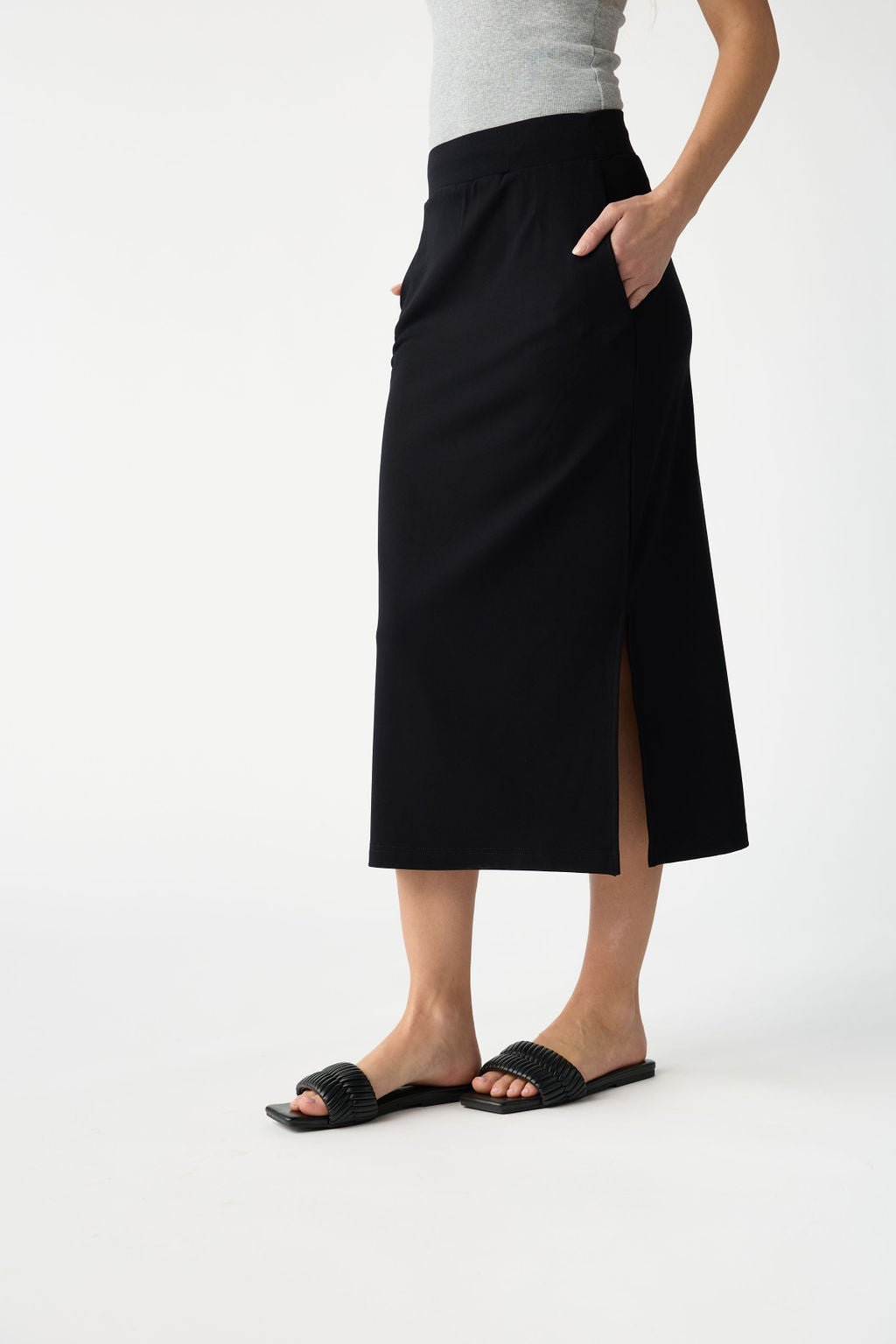 A person is wearing a gray sleeveless top, Cozy Earth's black Women's Brushed Bamboo Midi Skirt with a side slit, and black open-toe sandals. They stand with hands in pockets against a plain white background.