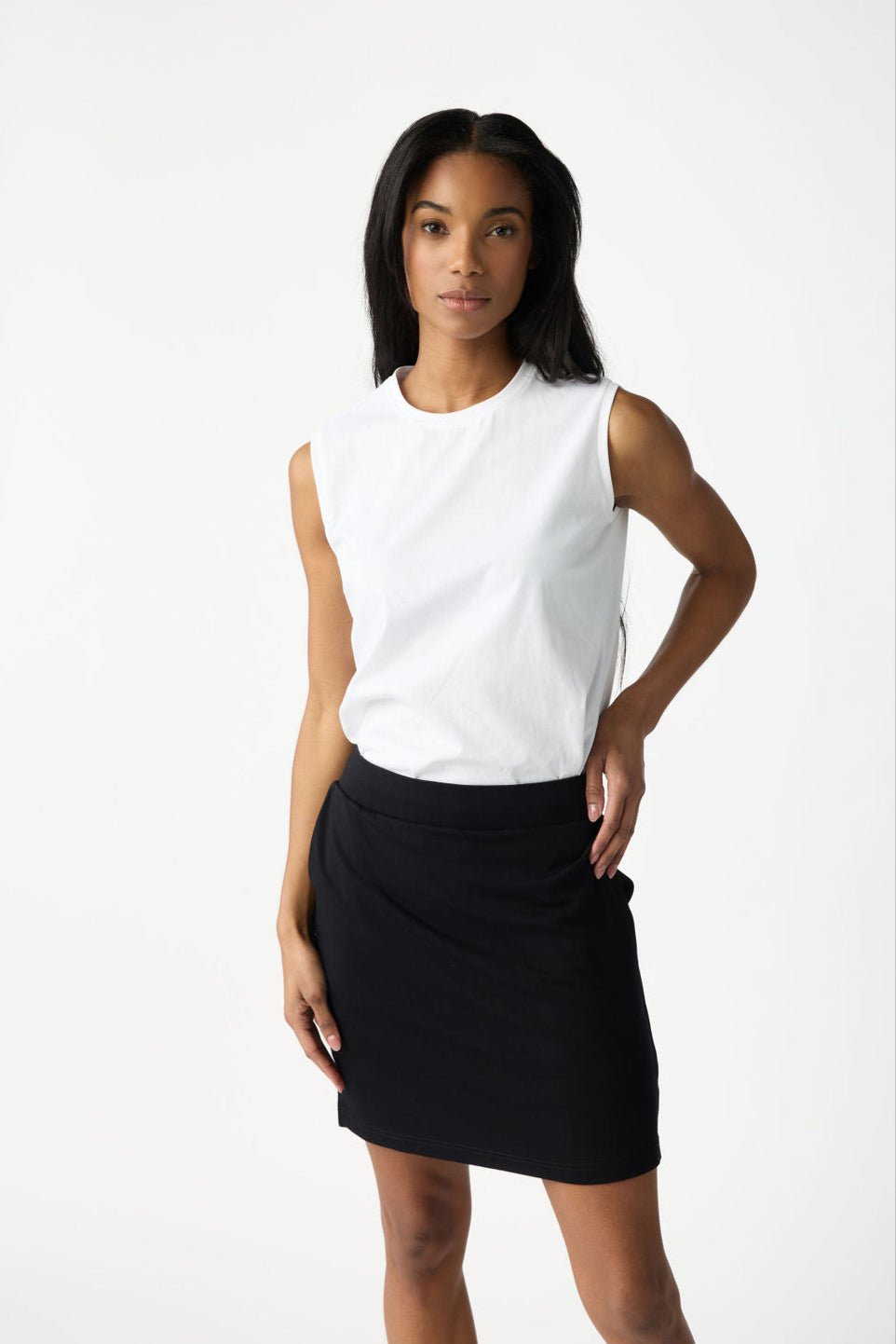 A person with long dark hair poses against a plain white background, wearing a sleeveless white top and the Women's Brushed Bamboo Skirt by Cozy Earth. Their right hand rests on their hip, while the left hangs by their side. 