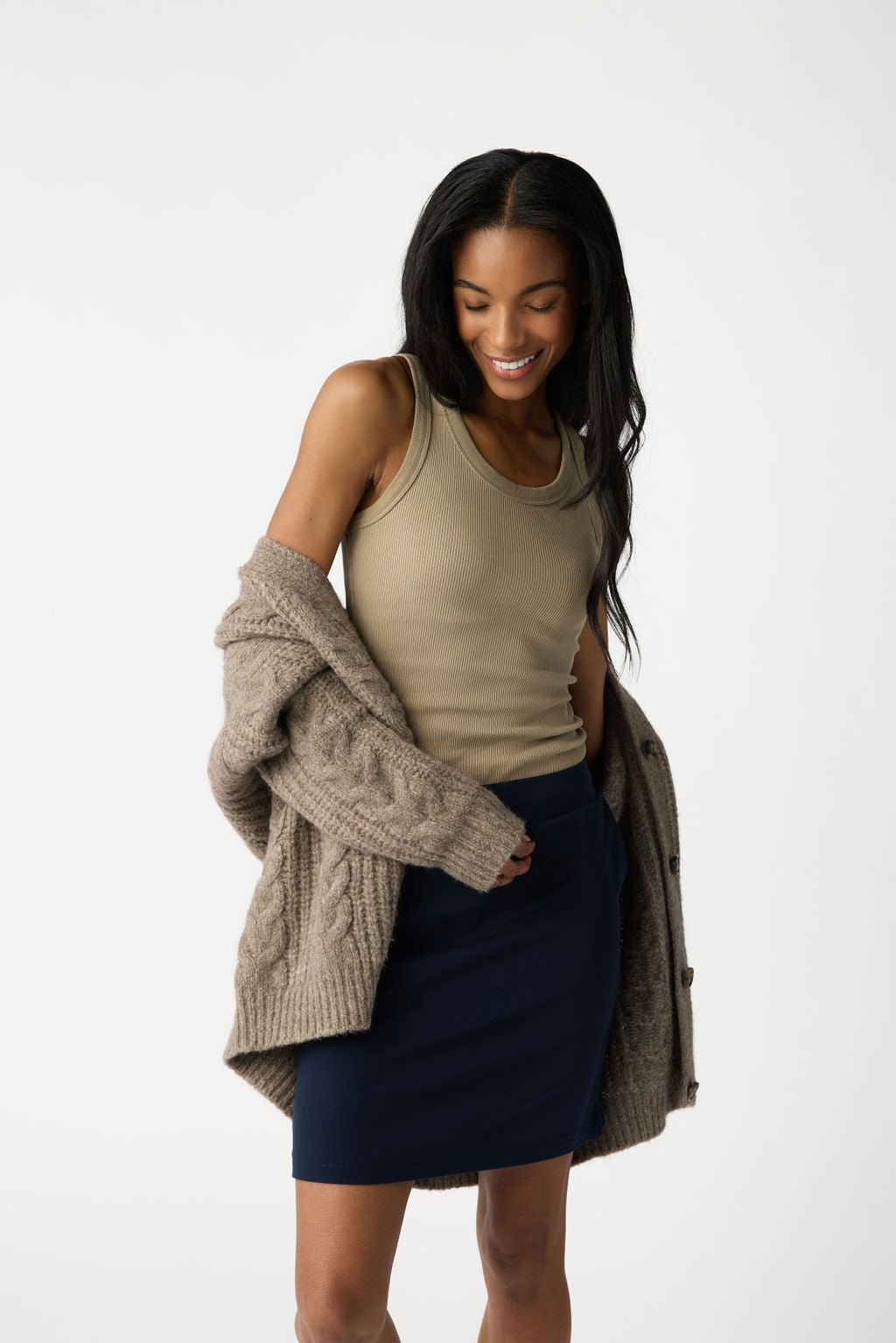 A woman smiles in Cozy Earth's Women's Brushed Bamboo Skirt with a beige tank top and a knitted cardigan draped over her shoulders. The background is plain white. 