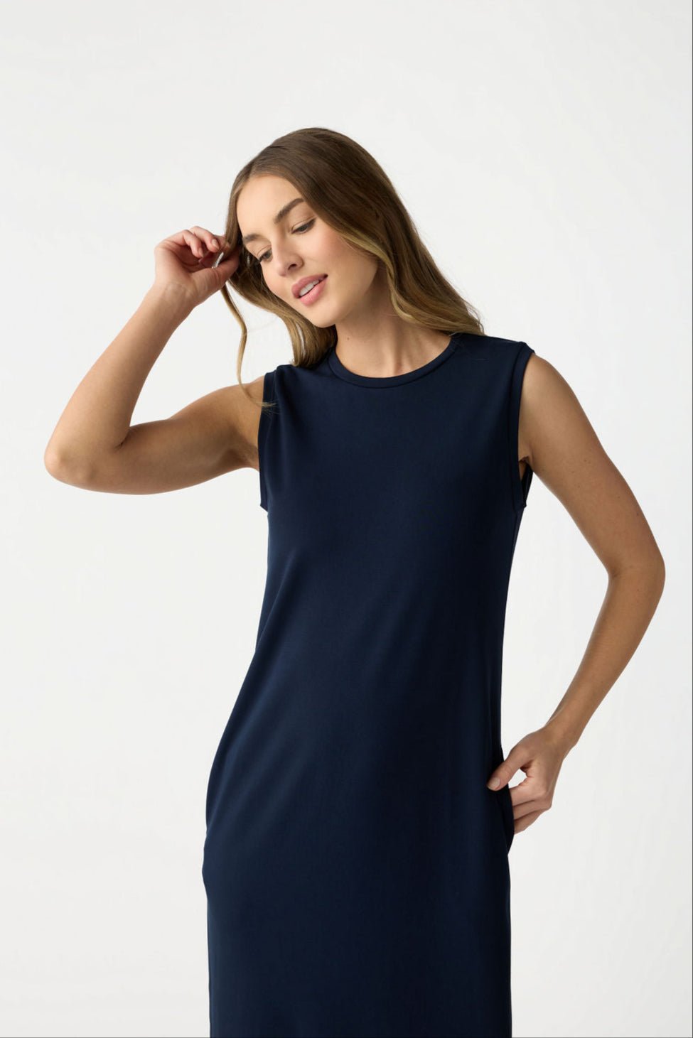 A woman with long wavy hair models the Cozy Earth Women's Brushed Bamboo Sleeveless Midi Dress in navy, striking a pose with one hand on her hip and the other gently touching her head against a light-colored background. 