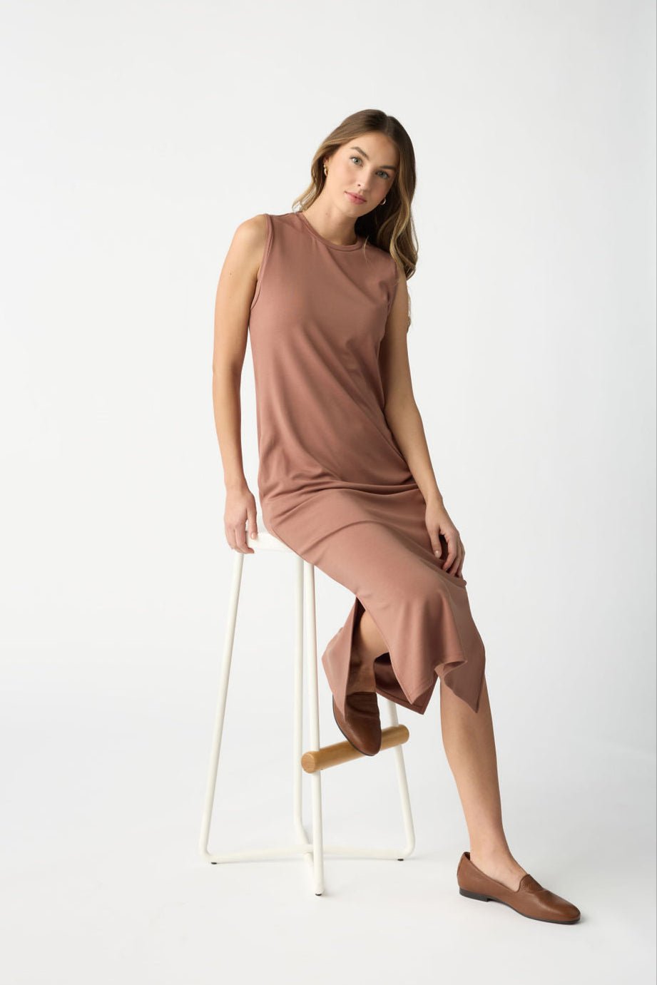 A woman in Cozy Earth's Women's Brushed Bamboo Sleeveless Midi Dress, in light brown, sits sideways on a white-framed stool with a brown wooden footrest. She has long hair and wears flat brown shoes against a plain white background. 