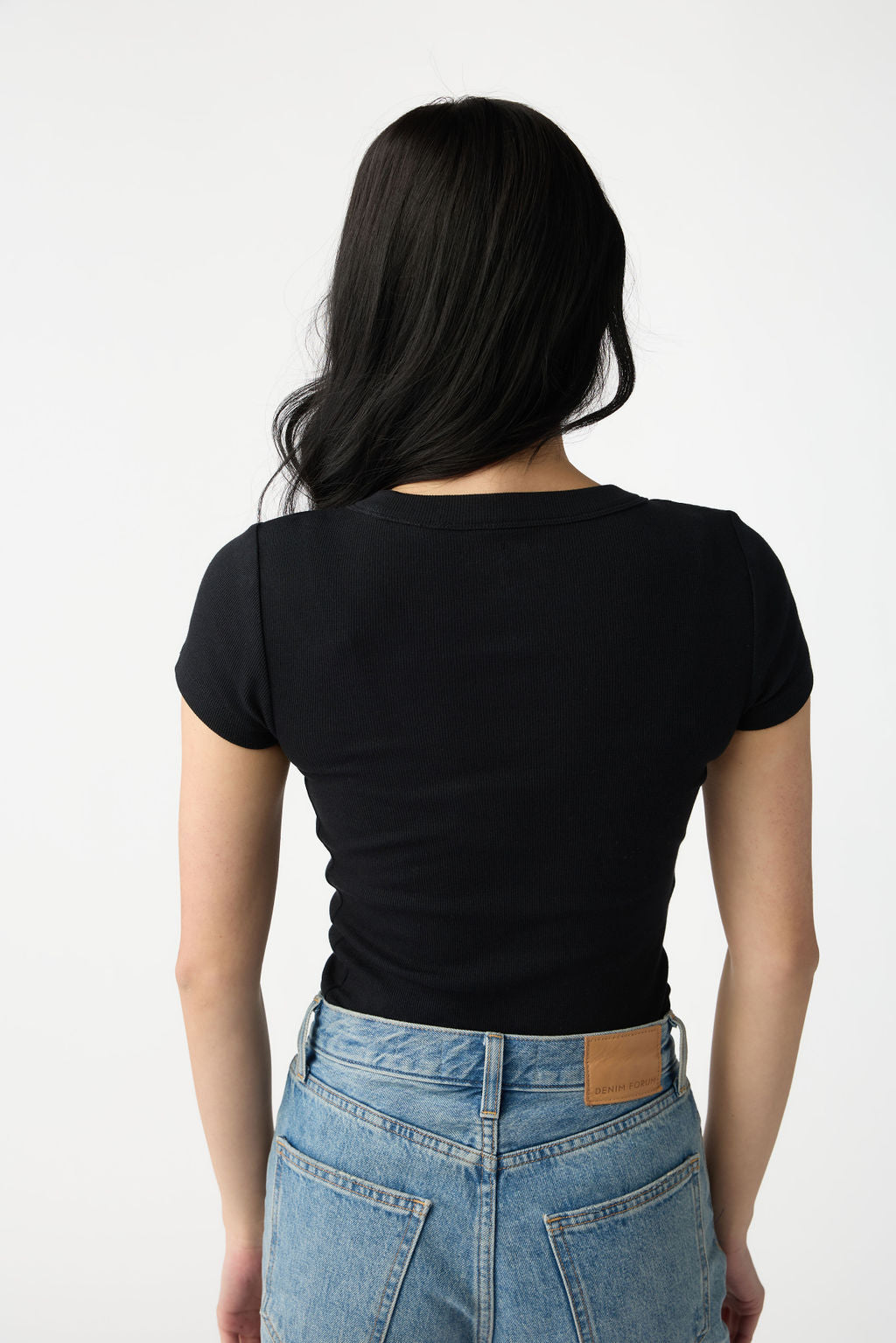 A dark haired woman wears Cozy Earth Women's Fine Ribbed Tee in Jet Black. She is facing away from the camera and stands in front of a white background. 