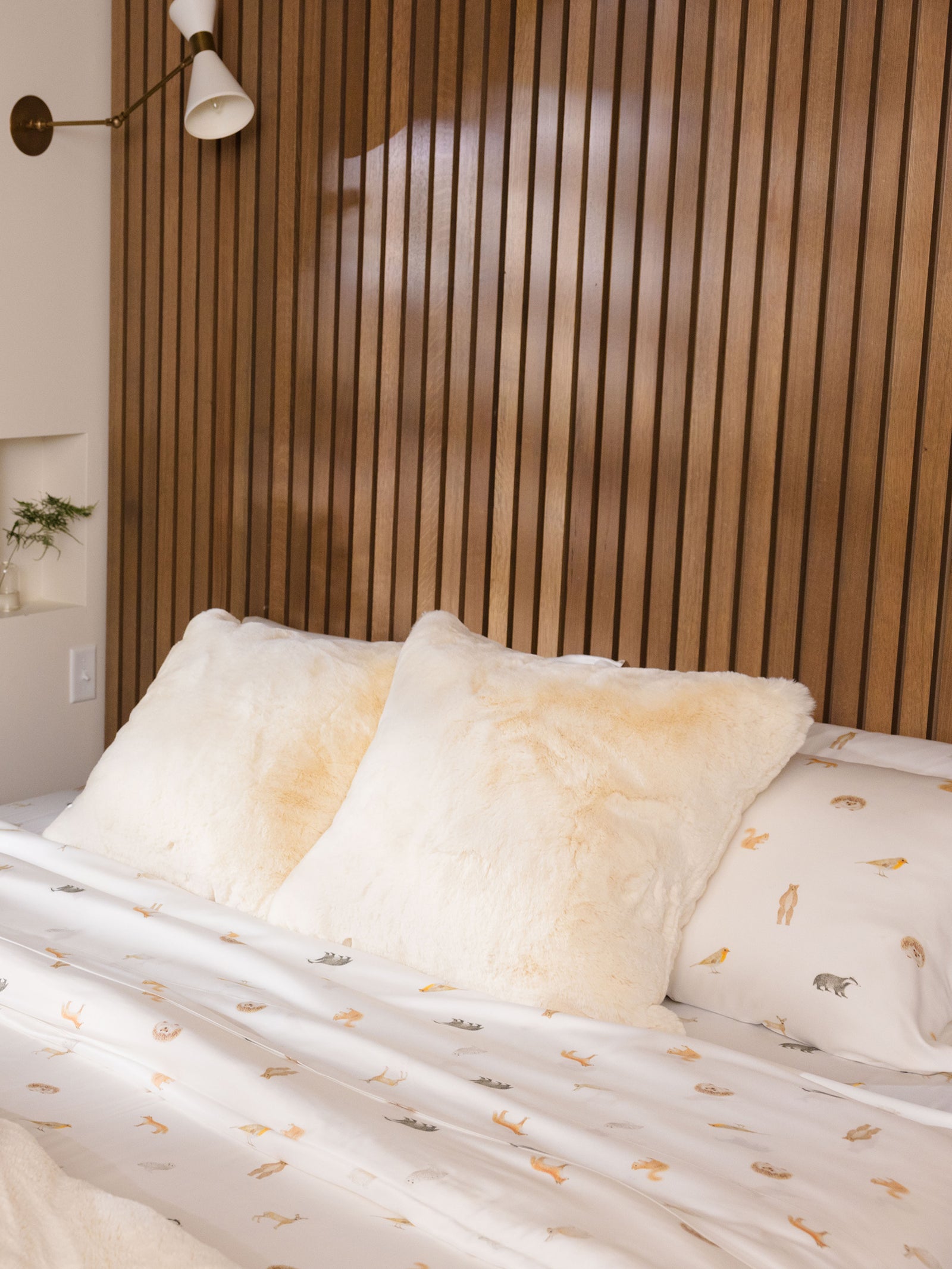 A cozy bed featuring Cozy Earth's Bamboo Duvet Cover, complemented by two large, fluffy cream-colored pillows against a wooden slat headboard. The bedspread boasts a minimalist illustration print, and there's a wall-mounted white lamp on the left side. 