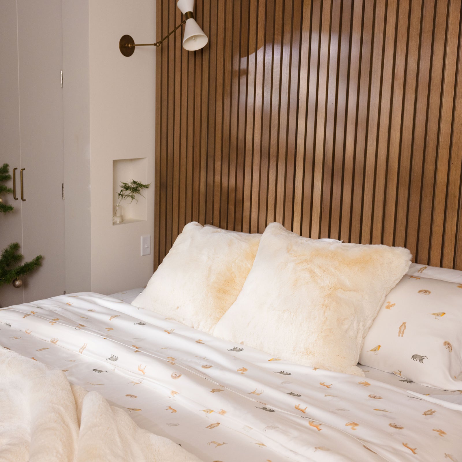 A cozy bedroom features a bed covered with the Bamboo Sheet Set by Cozy Earth, showcasing white sheets adorned with small animal prints and topped with fluffy white pillows. The wall behind the bed boasts vertical wooden paneling, complemented by a mounted white wall lamp above. Nearby, a small plant rests on a recessed shelf. 