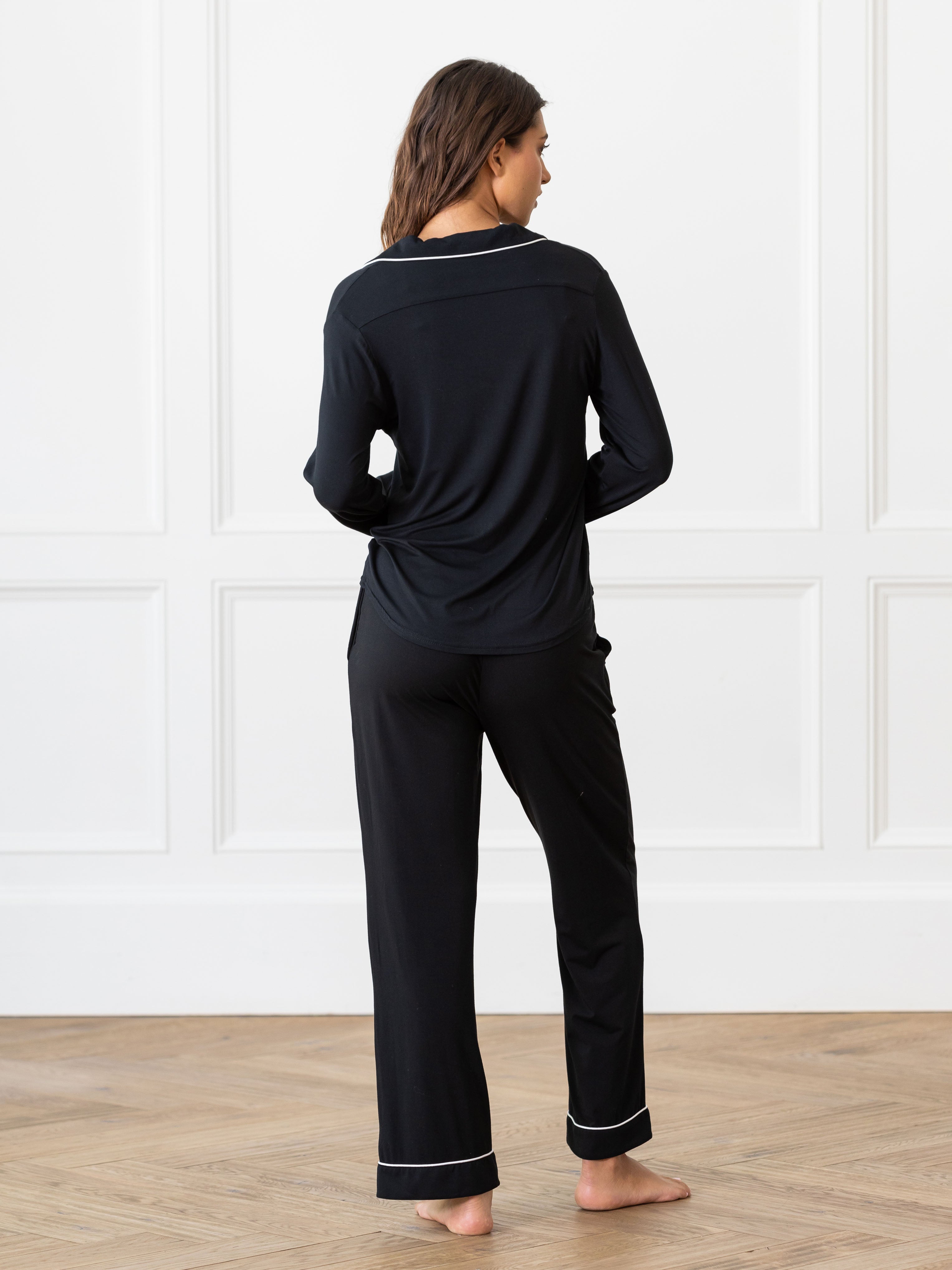 A woman with long brown hair, dressed in Cozy Earth's Women's Bedtime Bundle black pajamas, stands barefoot with her back facing the camera. The pajamas include a long-sleeve top and pants, both accented with white piping along the edges. She stands on a wooden floor against a white paneled wall background. |Color:Black