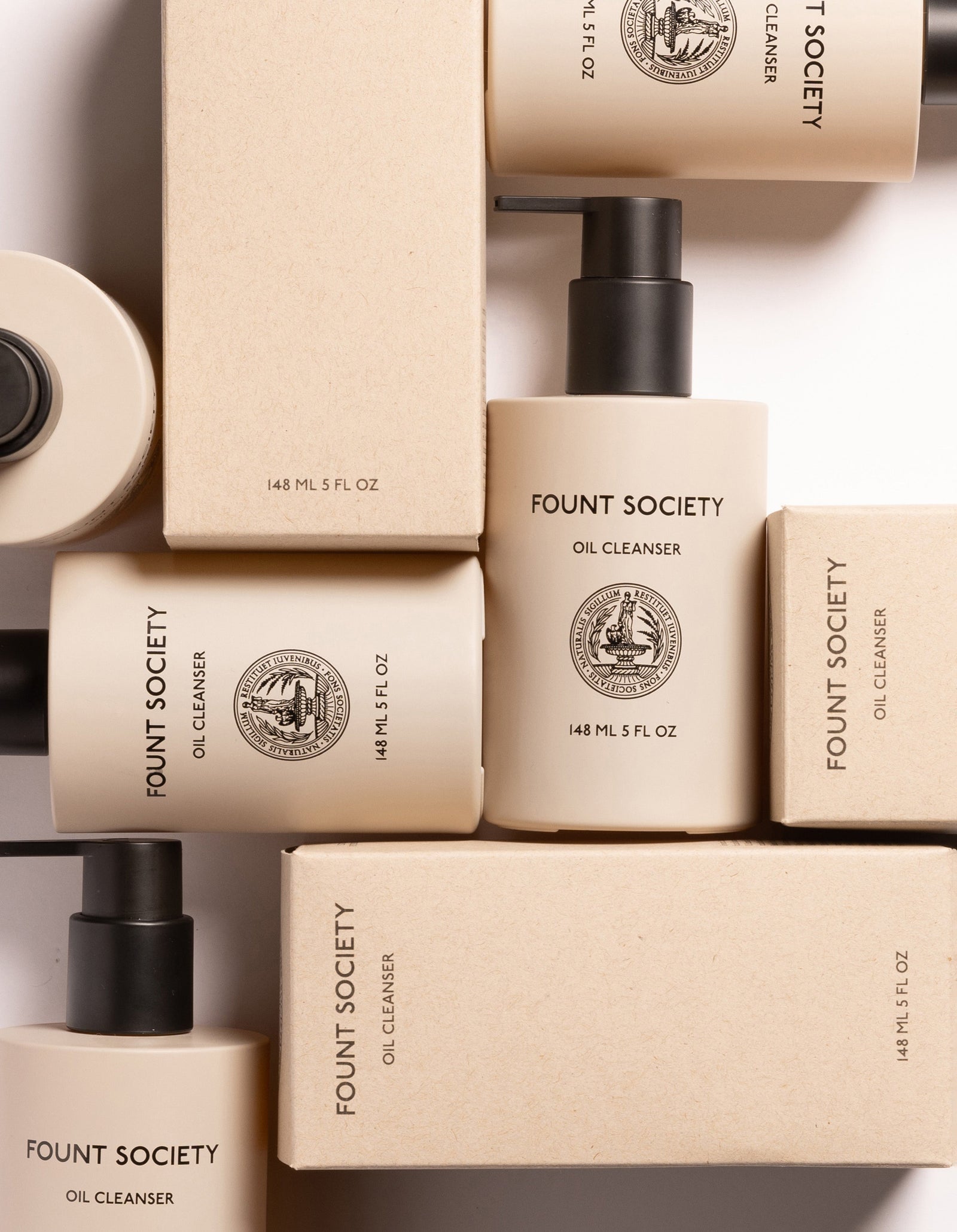 Flat lay of bottles and boxes of Cozy Earth Oil Cleanser, featuring a minimalistic design in a beige color palette with a black pump dispenser. The product details and brand logo are visible on both the bottles and packaging.