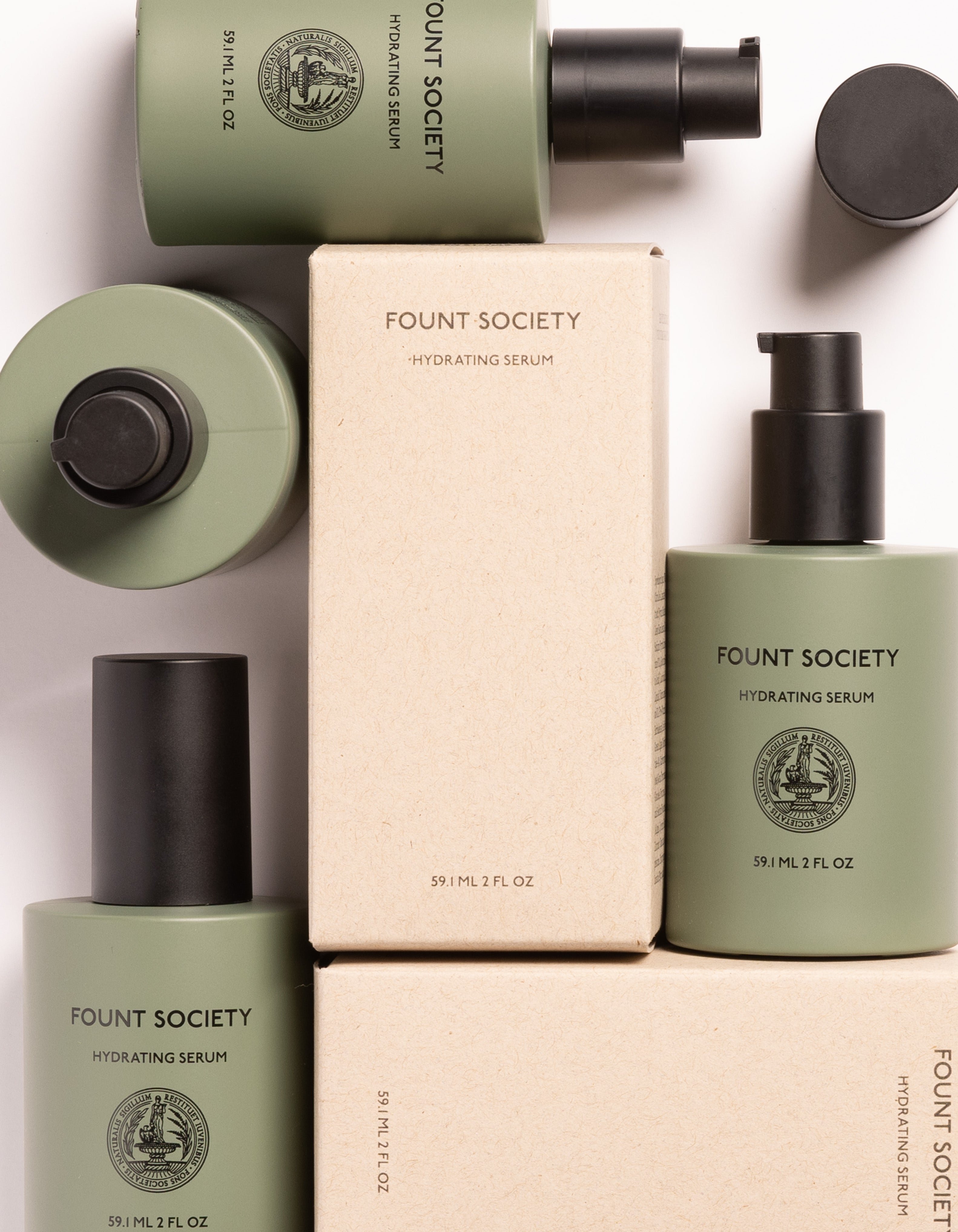 A flat lay photo of Cozy Earth Hydrating Serum products. The image features green bottles with black pumps and a matching green cap, alongside cream-colored boxes. Each bottle is labeled with "Cozy Earth Hydrating Serum 59 mL / 2 fl oz".