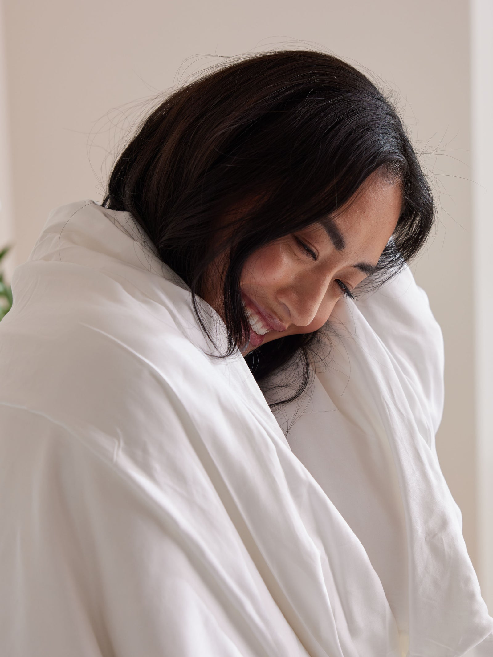 A person with long dark hair smiles softly while wrapped in a Cozy Earth Silk Comforter, looking relaxed and content in a softly lit room.