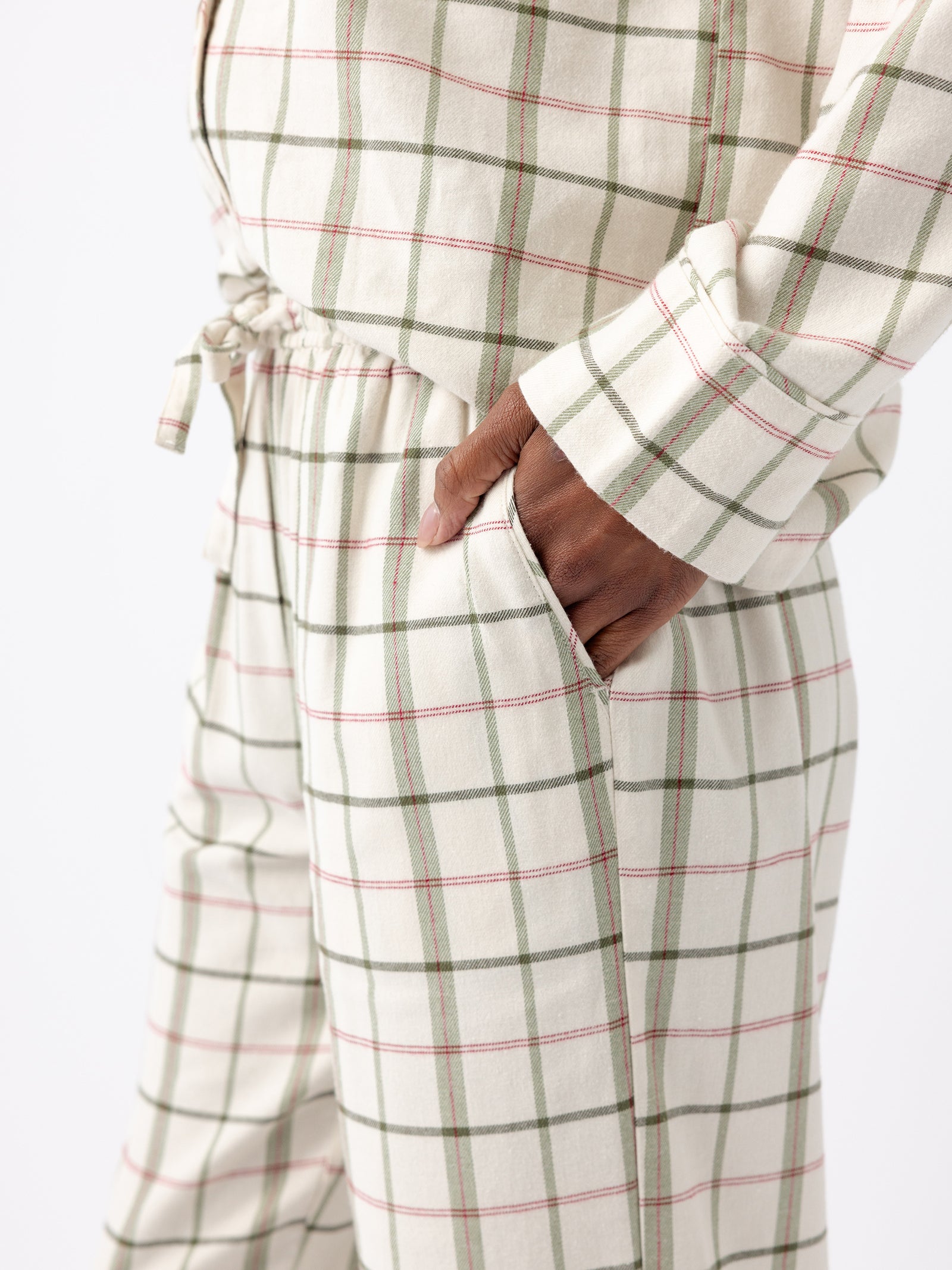 A person wearing the Women's Farmhouse Pajama Pant from Cozy Earth, characterized by its plaid pattern in beige, green, and pink. The set includes a long-sleeve shirt and pants with an adjustable drawstring waist. The person has one hand inside the pant pocket. 