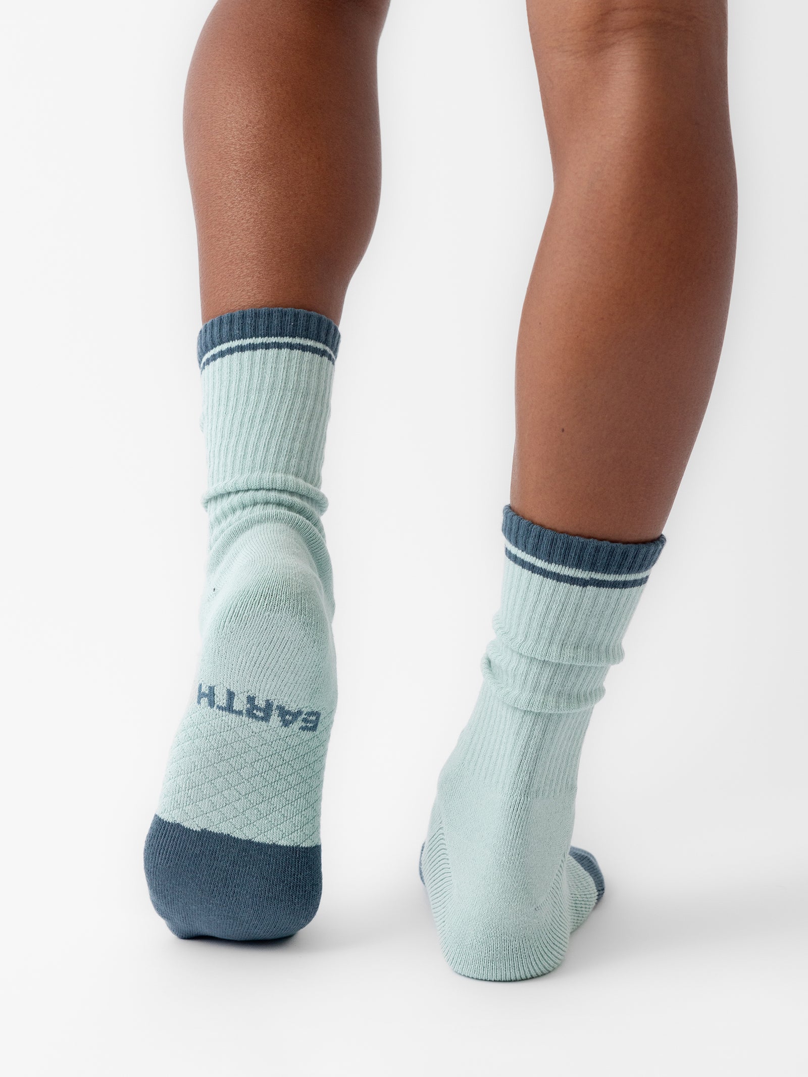 A person is wearing Cozy Earth’s Essential Calf Socks in light green, featuring dark green accents at the cuffs, toes, and heels. The word "EARTH" is printed on the bottom of one of the socks. The person is standing with their back to the camera, revealing the back of their legs and feet. 