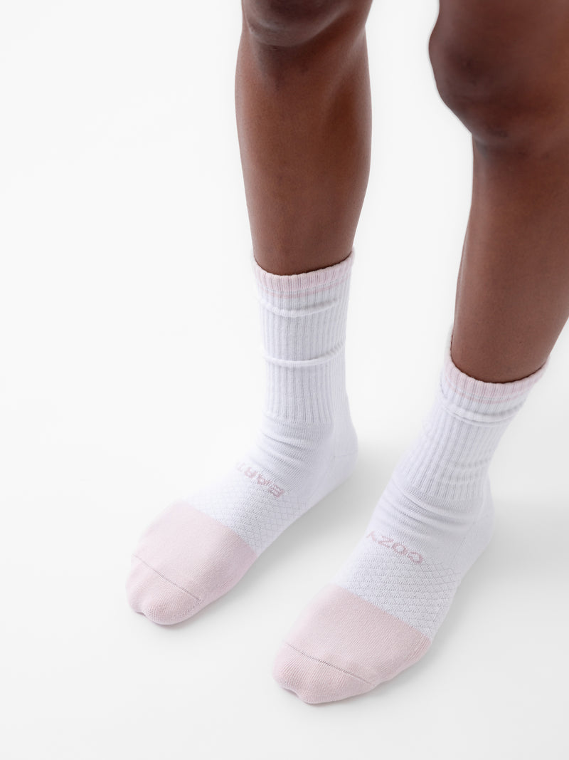 A close-up shot features a person donning the white ribbed socks from Cozy Earth's Essential Calf Sock 4-Pack. The socks showcase light pink toes and have "COZY" emblazoned on one and "SMART" on the other, all against a plain white backdrop.