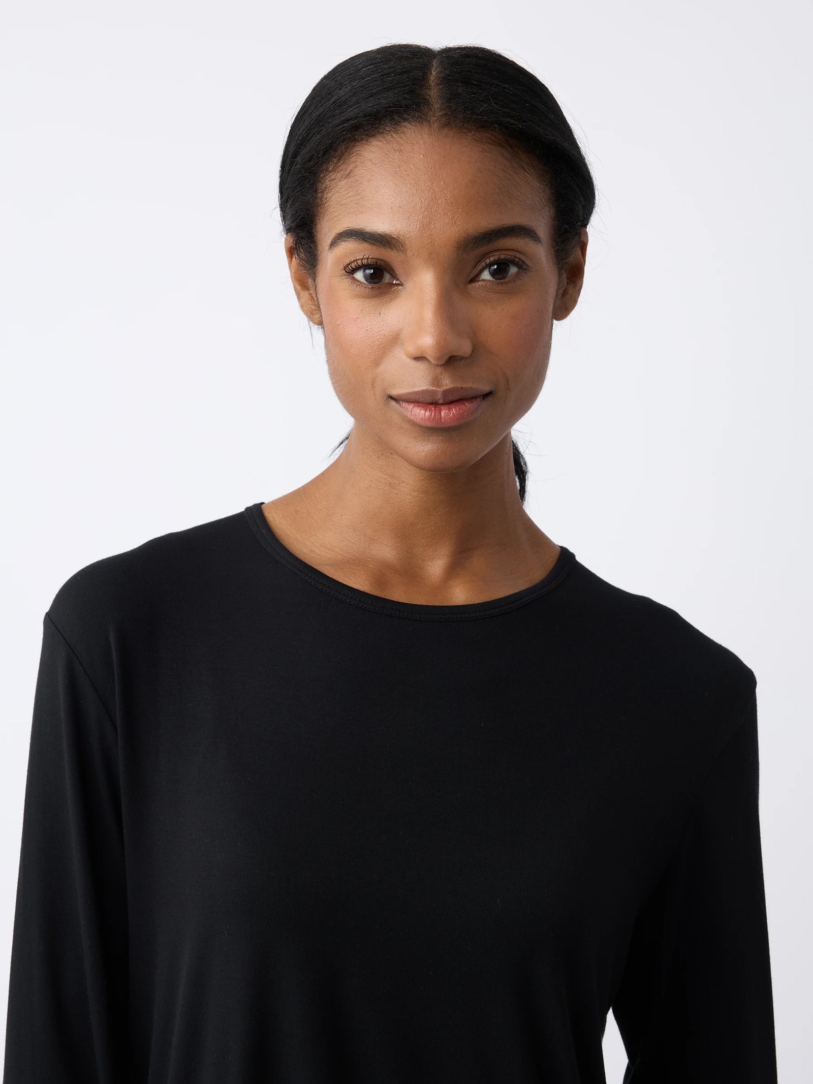 A woman with long dark hair is wearing a Women's Stretch Knit Bamboo Long Sleeve Lounge Tee from Cozy Earth, looking at the camera with a neutral expression against a plain, light-colored background. 