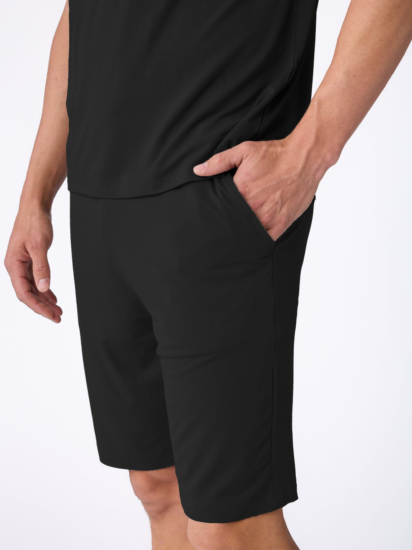 A man wearing the Cozy Earth Men's Bamboo Stretch-Knit Short Sleeve Pajama Set, featuring a black short-sleeve shirt and black knee-length shorts, stands against a plain white background with one hand in his pocket. 