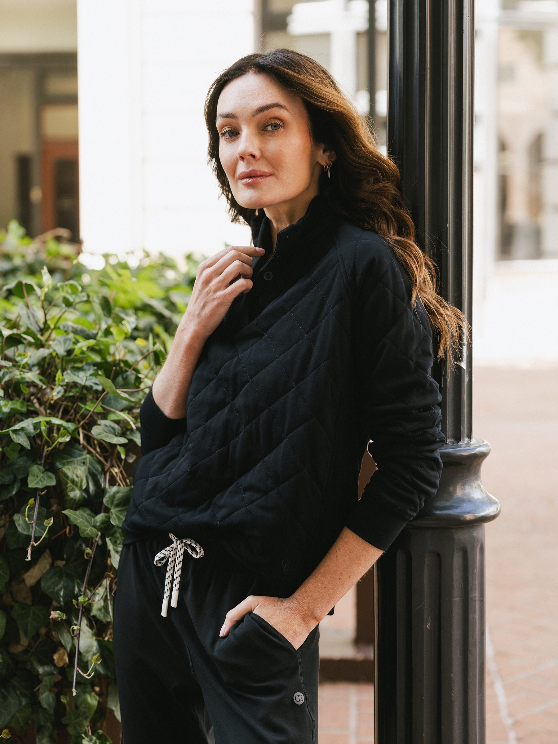 A woman with long, wavy hair leans against a lamppost outdoors. She is wearing Cozy Earth's Women's Ultra-Soft Bamboo Quilted Snap Pullover in black and black drawstring pants. Her right hand is near her neck, and her left hand is in her pocket. There are green plants and buildings in the background. 