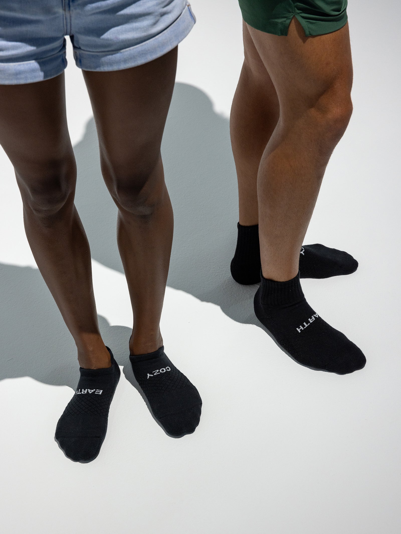 Two people are standing side by side on a light-colored floor. Both are wearing black socks from the Essential Ankle Sock 2-Pack by Cozy Earth, though the text on them is partially out of frame. One person is wearing green shorts and the other is in light blue shorts. 