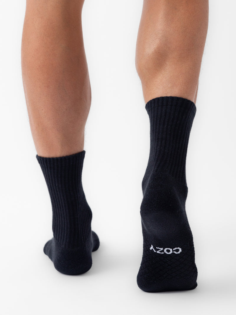 A person stands on tiptoe, viewed from behind, wearing black Essential Calf Socks by Cozy Earth. "COZY" is printed in white on the sole of the right sock. The background is plain white.