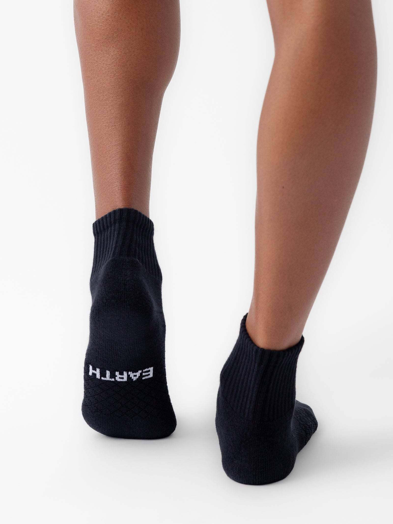 A person is wearing black ankle socks from the Essential Quarter Sock 2-Pack by Cozy Earth, featuring the word "EARTH" written in white on the sole. The image captures the back of their legs and feet against a plain white background. 