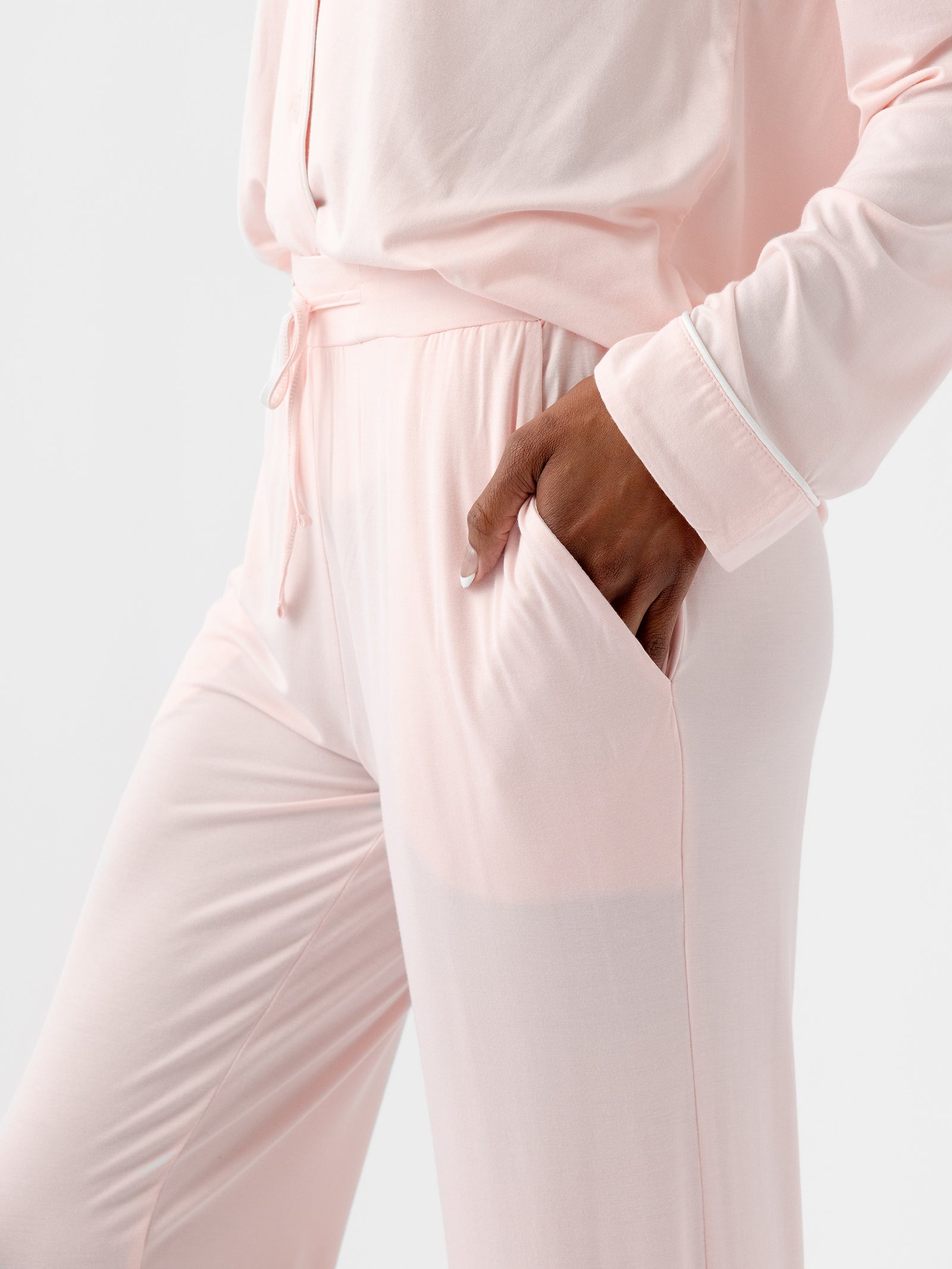 A person dressed in HIDE's Women's Stretch-Knit Bamboo Pajama Pant TALL in light pink is shown from the shoulders down, with their left hand in the pocket of their pants. The soft and loose-fitting outfit features a long-sleeve top and drawstring pants against a plain white background. 