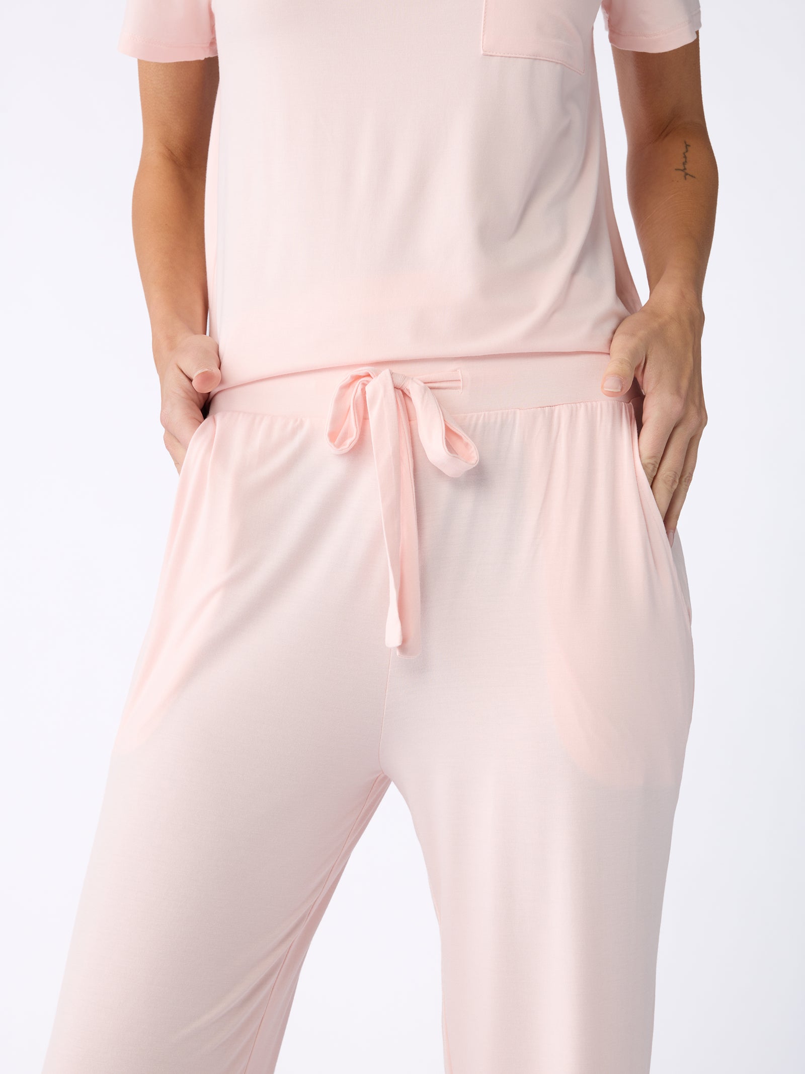 A person wearing a pale pink short-sleeved top paired with Cozy Earth's Stretch Knit Bamboo Pant, featuring a drawstring, is standing with their hands in the pockets against a light background. 