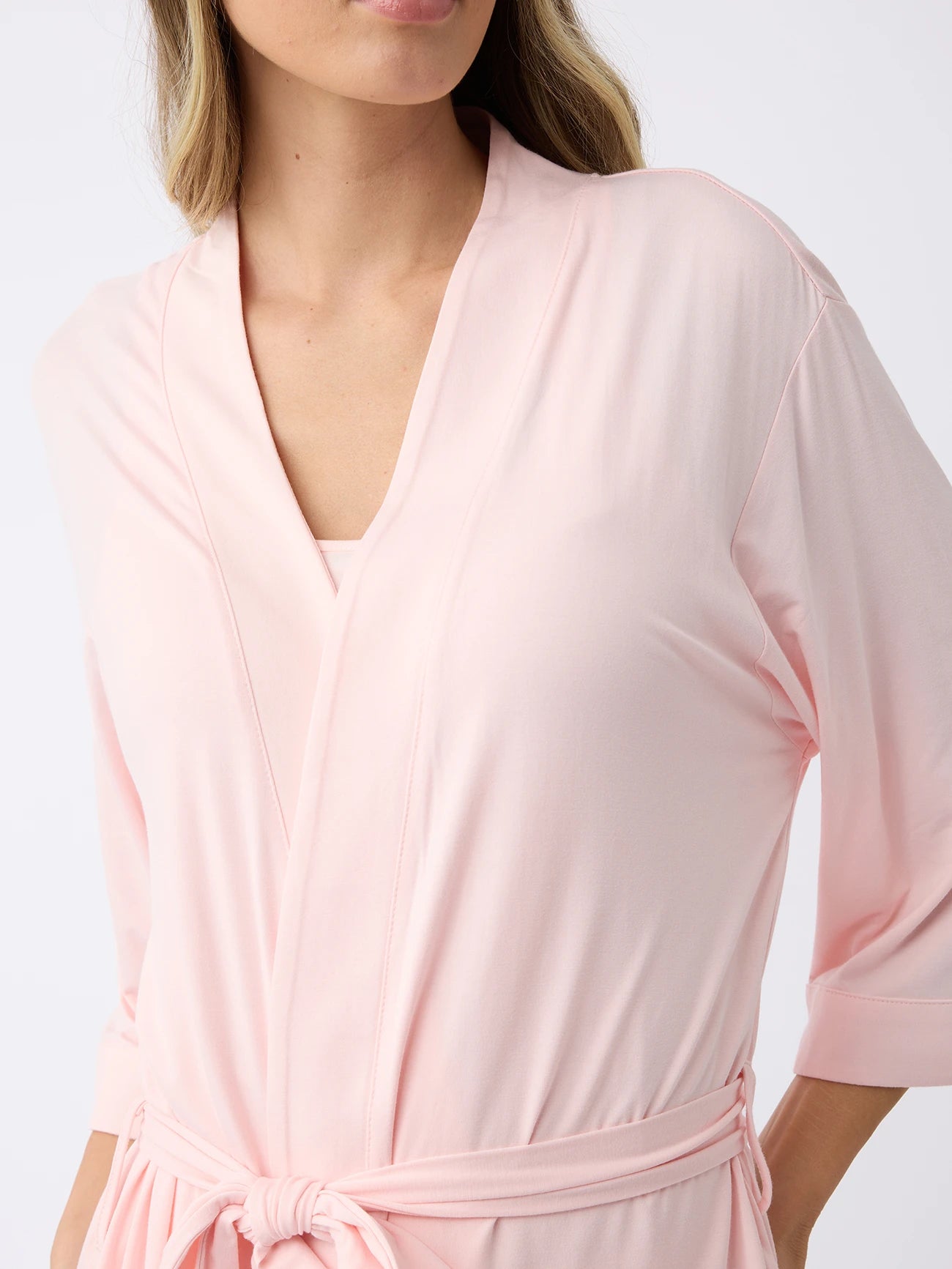 A person wearing Cozy Earth's Women's Stretch-Knit Bamboo Kimono Robe in light pink, featuring a tied belt and three-quarter sleeves. The plain white background accentuates the robe's soft fabric and relaxed fit, with only the upper body and part of the face visible. 