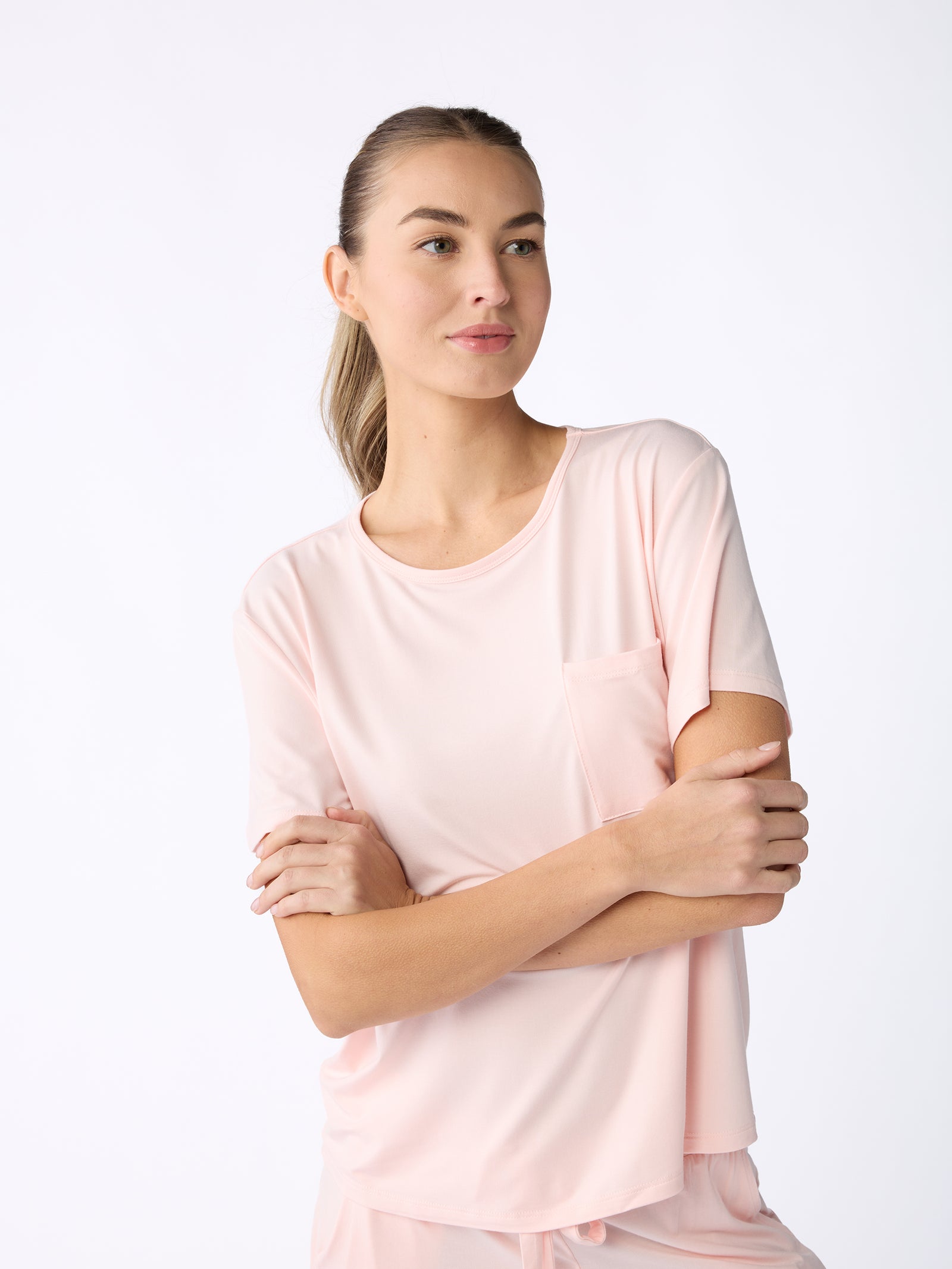 A person is wearing Cozy Earth's Women's Stretch-Knit Bamboo Lounge Tee in light pink, featuring short sleeves and a pocket, standing with arms crossed against a plain white background. 