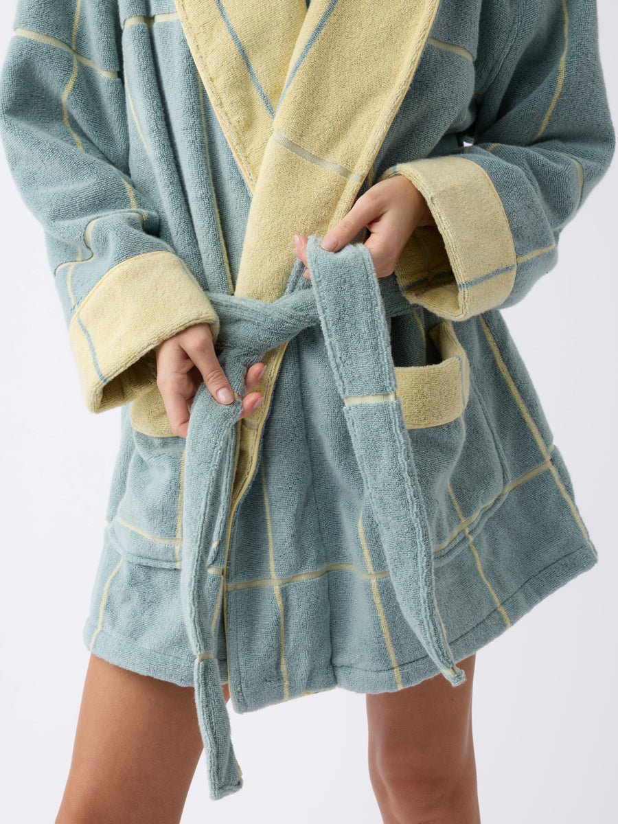 A person is tying the belt of the Cozy Earth Windowpane Resort Robe, which is light blue with a yellow grid pattern and cuffs, while facing forward. Only their hands and partially visible legs can be seen. 