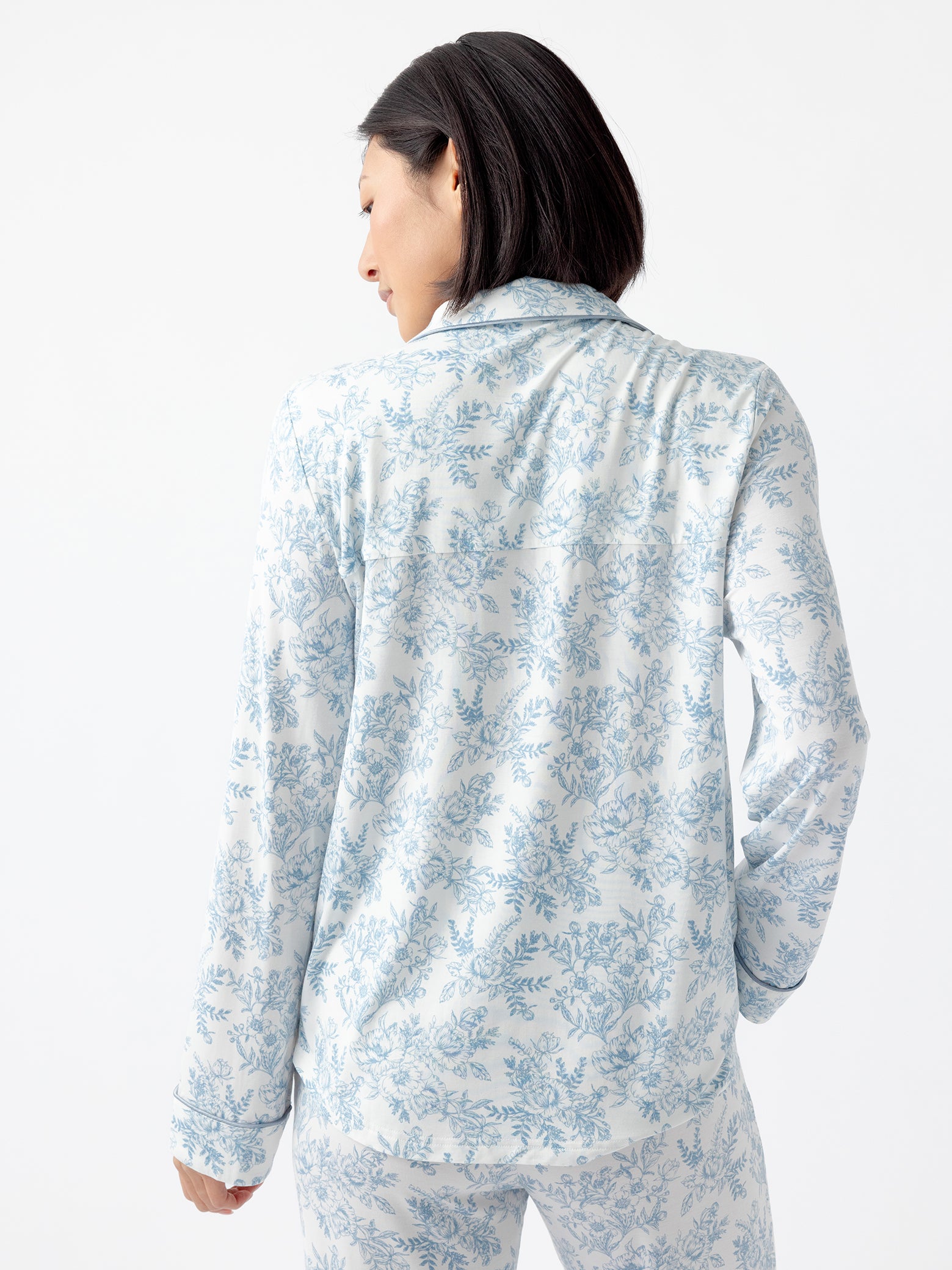 A person with short dark hair faces away from the camera, wearing the Cozy Earth Women's Bamboo Stretch-Knit Long Sleeve Pajama Top with a light blue floral pattern. The background is plain and light-colored. 