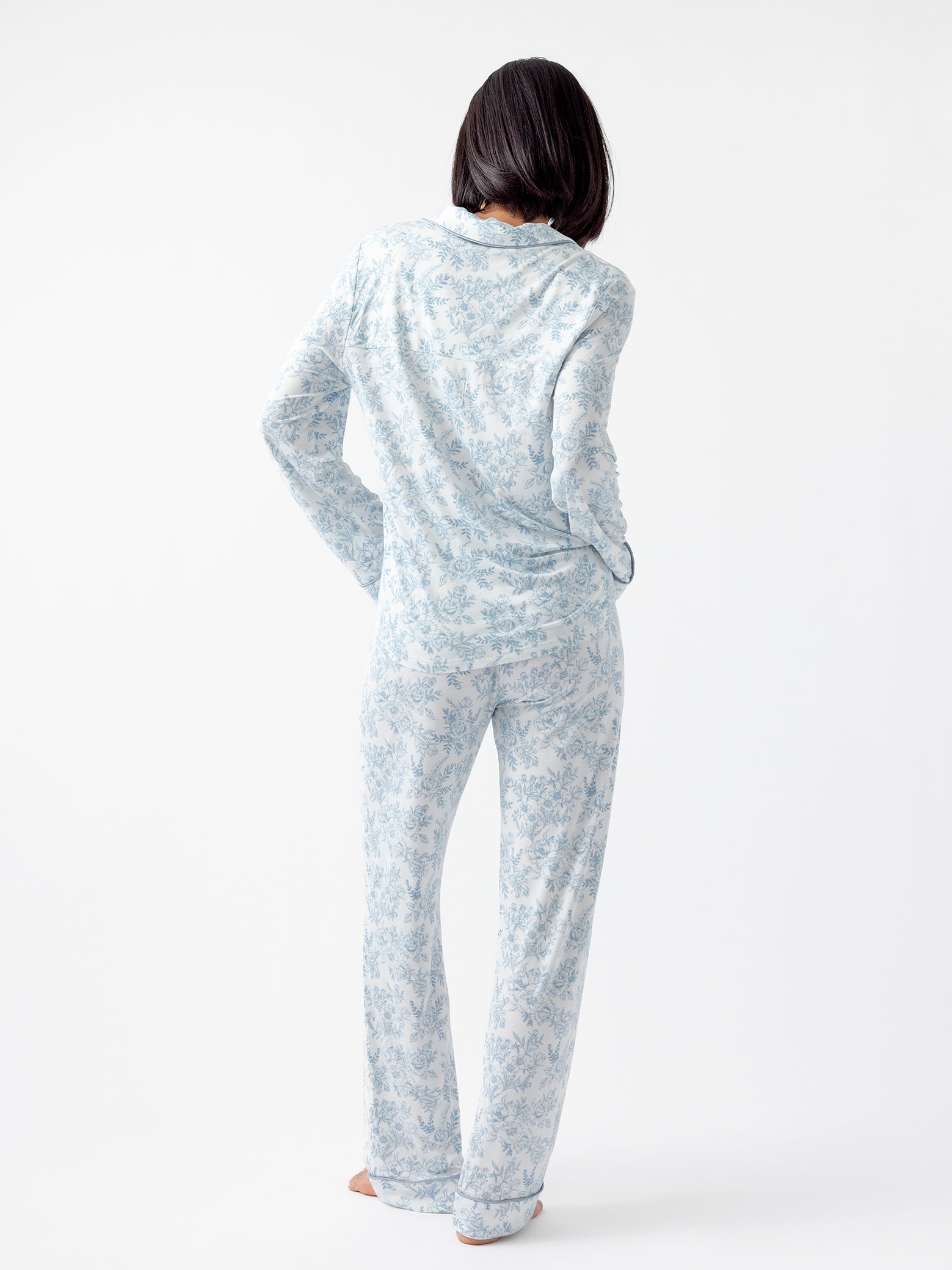 A barefoot individual with shoulder-length black hair is standing with their back to the camera, wearing a light blue floral pajama set featuring Cozy Earth Women's Bamboo Stretch-Knit Pajama Pant and a matching long-sleeve top against a plain white background. 