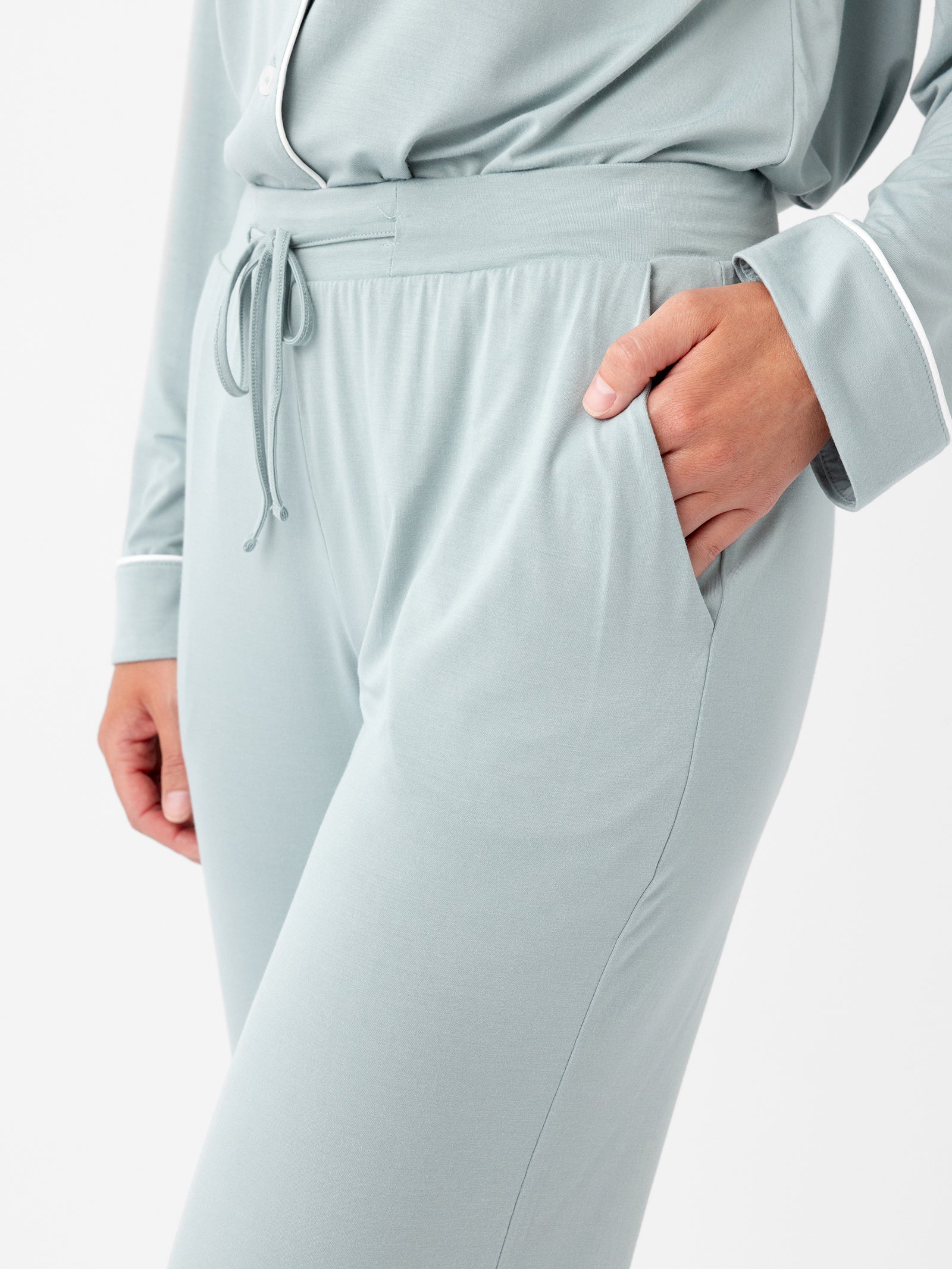 A person wearing the Cozy Earth Women's Stretch-Knit Bamboo Pajama Pant in light gray, featuring a drawstring waist, and a matching long-sleeved top adorned with white trim detail. The person has one hand in their pocket, highlighting the relaxed fit and soft fabric of this sleepwear ensemble. 