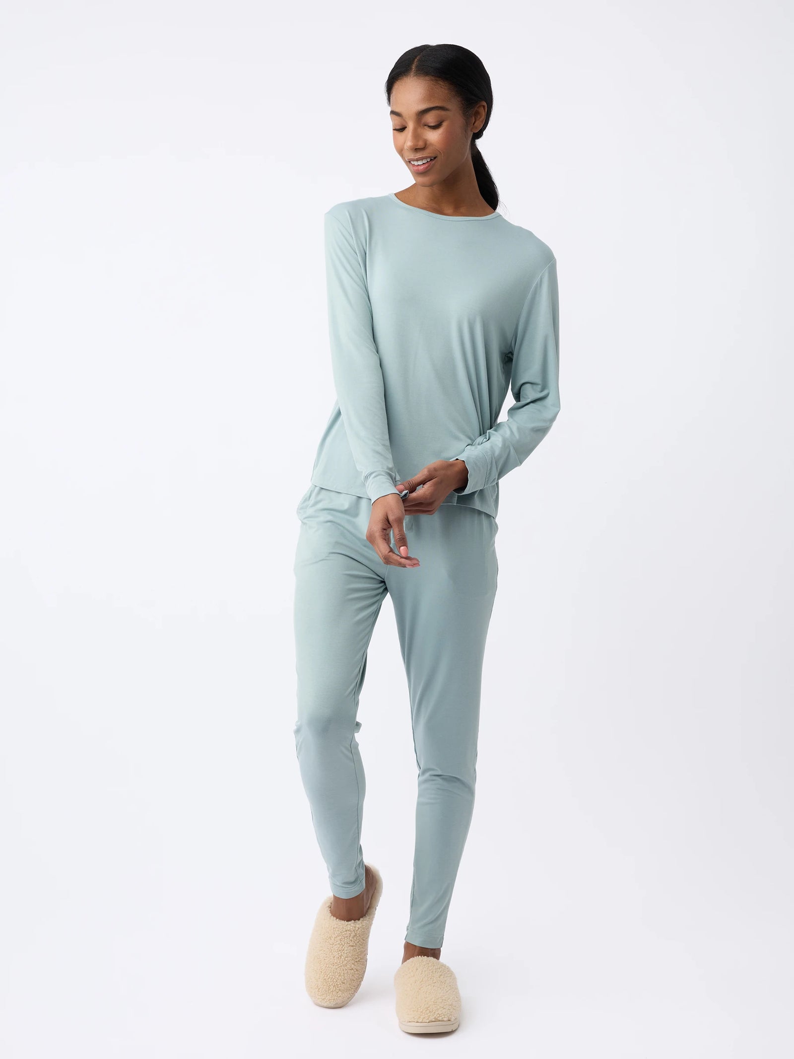 A woman in a light blue Cozy Earth Women's Stretch Knit Bamboo Long Sleeve Lounge Tee and fuzzy slippers stands against a plain white background, smiling and slightly looking downward. 