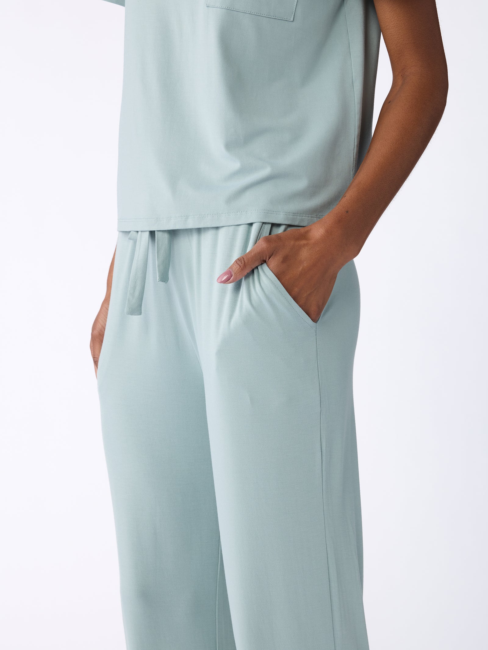 An individual is dressed in Cozy Earth's "Stretch Knit Bamboo Pant" in a light green shade, paired with a matching shirt. The ensemble features hands tucked into pockets, exuding a relaxed and casual vibe against a white background. 