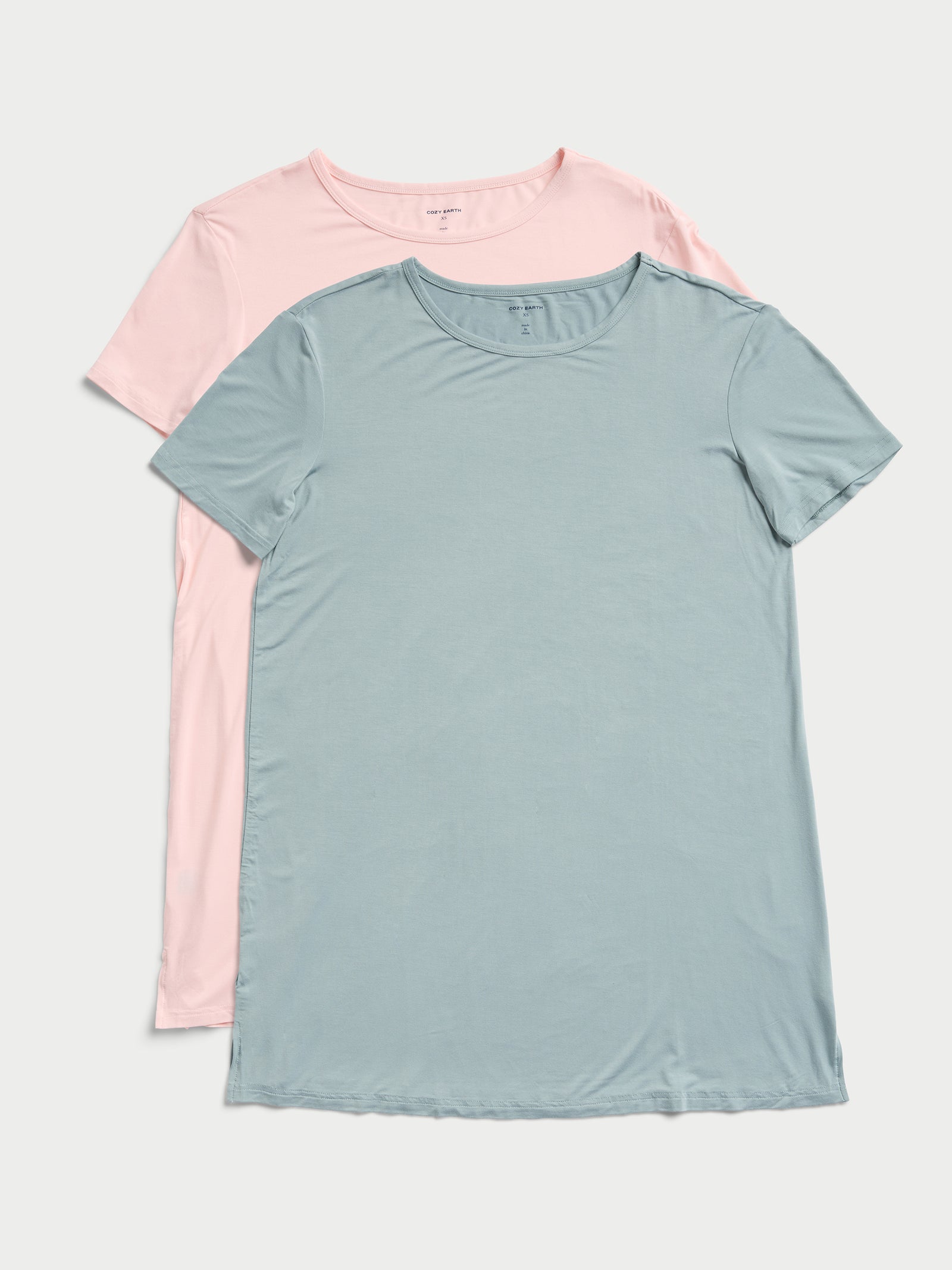 Two Women's Bamboo Stretch Knit Sleep Dresses from Cozy Earth are displayed on a flat surface. The dress on top is light blue, while the one underneath is pale pink. Both dresses feature short sleeves and round necklines. 