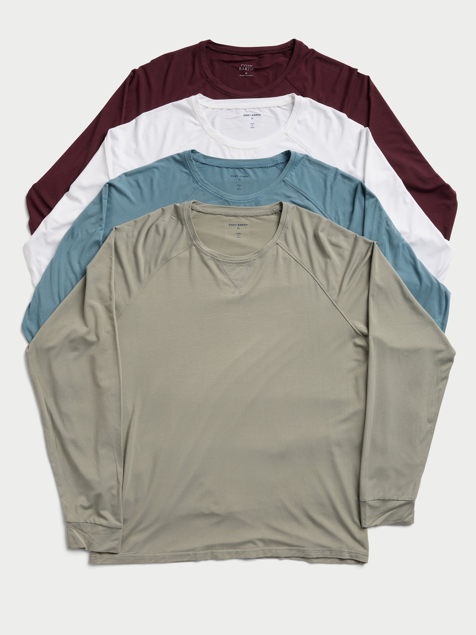 Four Men's Stretch-Knit Bamboo Long Sleeve shirts by Cozy Earth are neatly stacked. From top to bottom, the shirts come in maroon, white, teal, and olive green colors. They are laid flat, highlighting their simple crew neck design. 